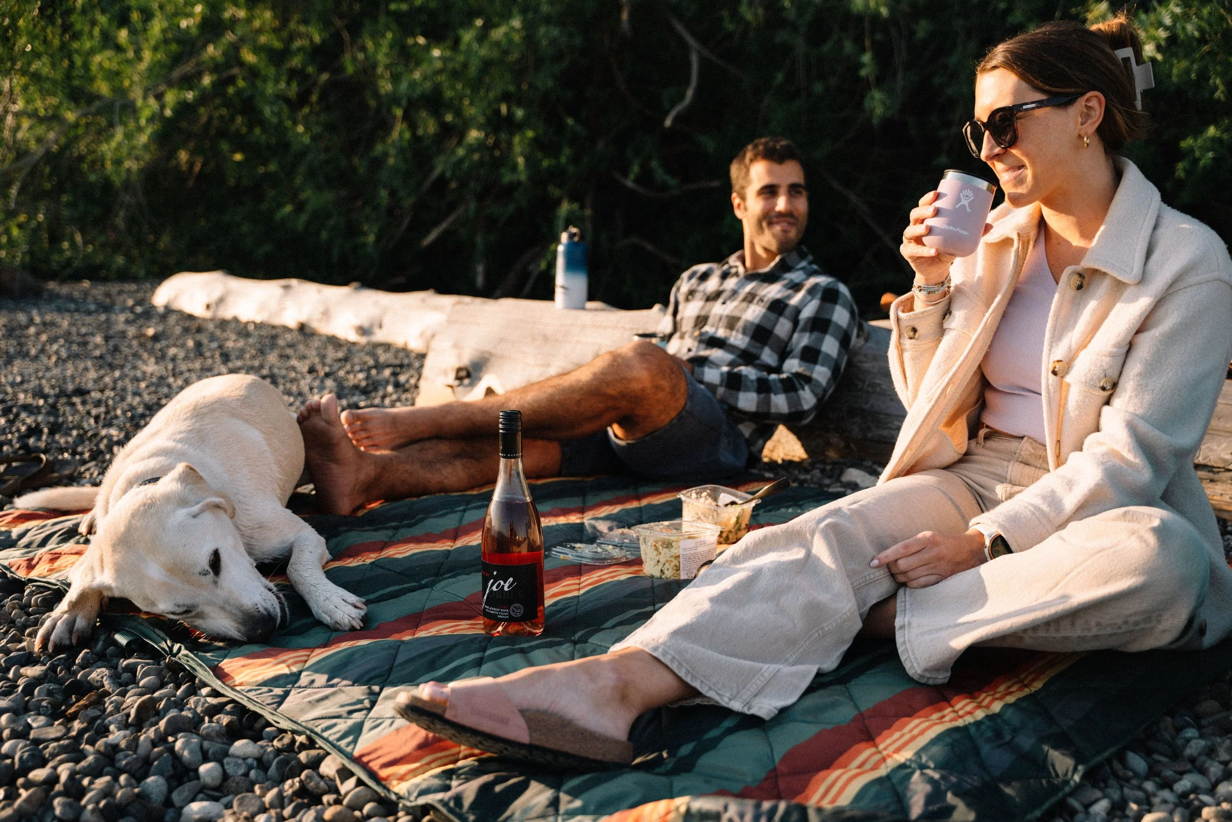 A woman and a man relaxing outdoors on a rocky beach, with a Labrador laying on a blanket, enjoying drinks and snacks during sunset.
