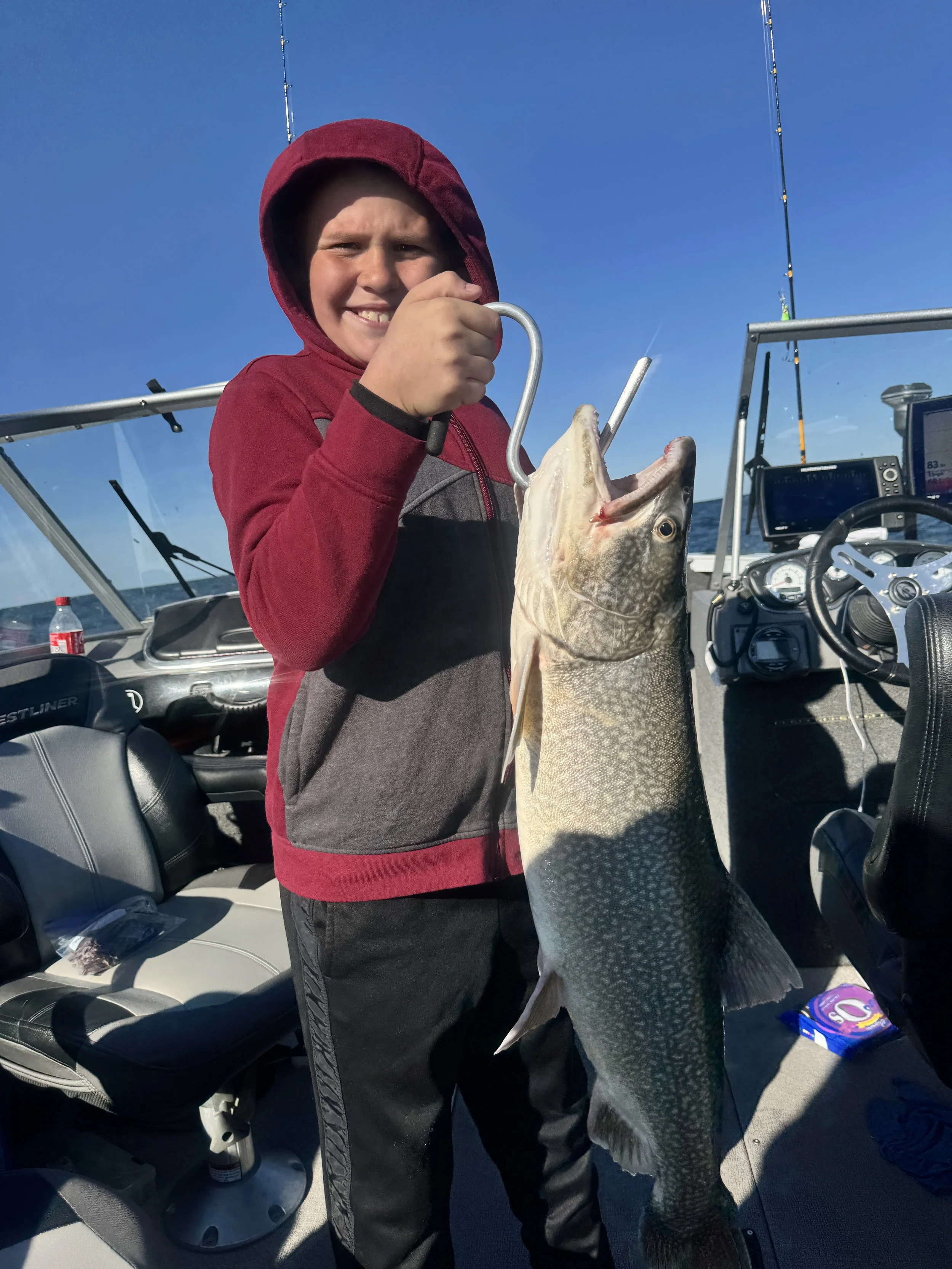 Young boy in a red hoodie holding a large fish on a boat with fishing gear and ocean in the background.