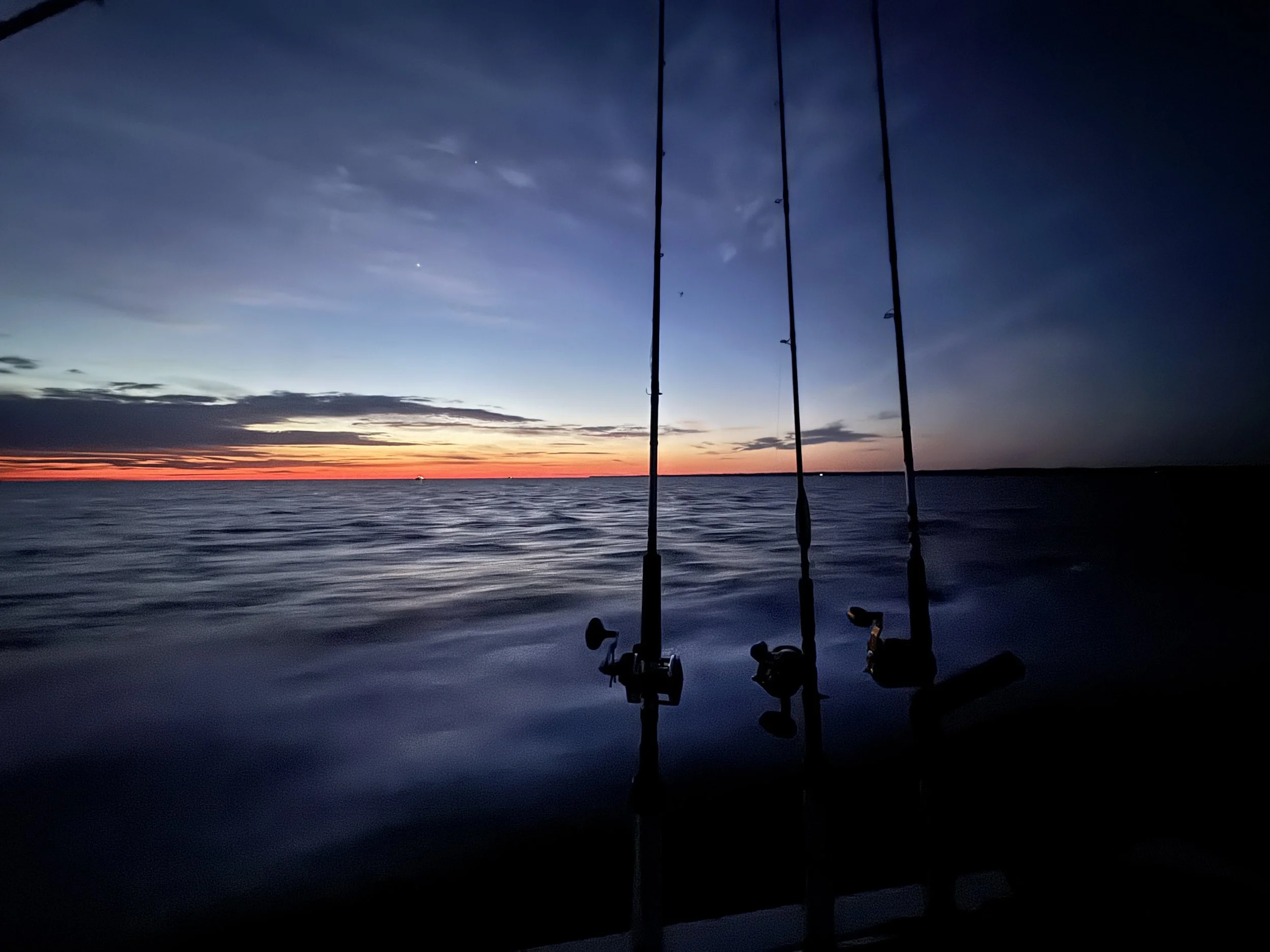 Three fishing rods are set up on a boat at sunset over calm water, with a colorful sky and distant horizon.
