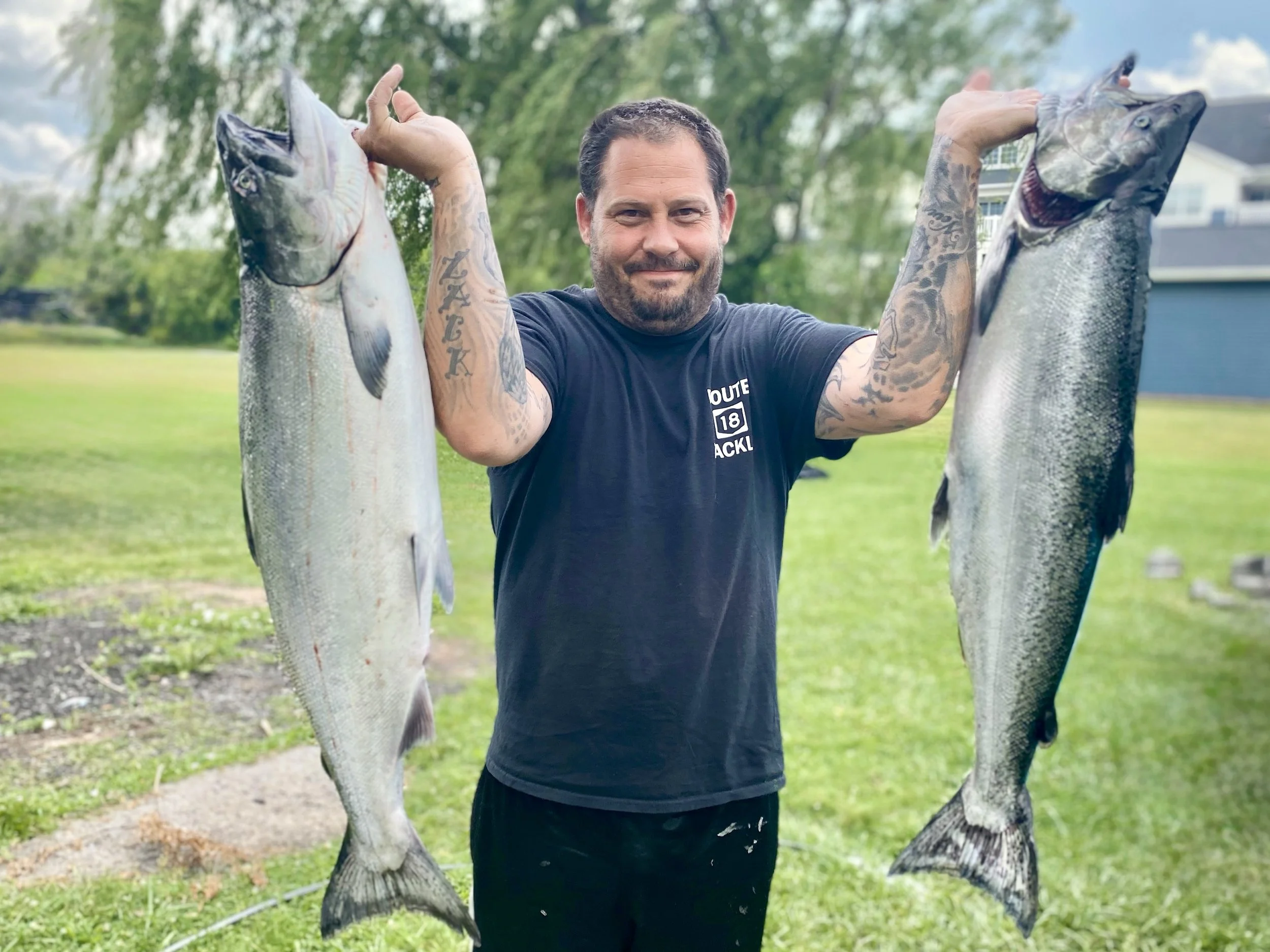 A man holding two large fish, one in each hand, outdoors on a grassy area with trees and houses in the background.