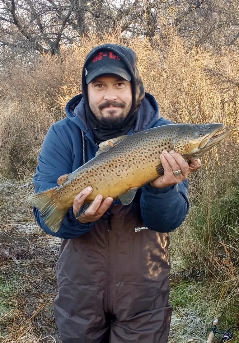 Man in outdoor clothing holding a large fish, standing near a bushy, marshy area with winter trees in the background.