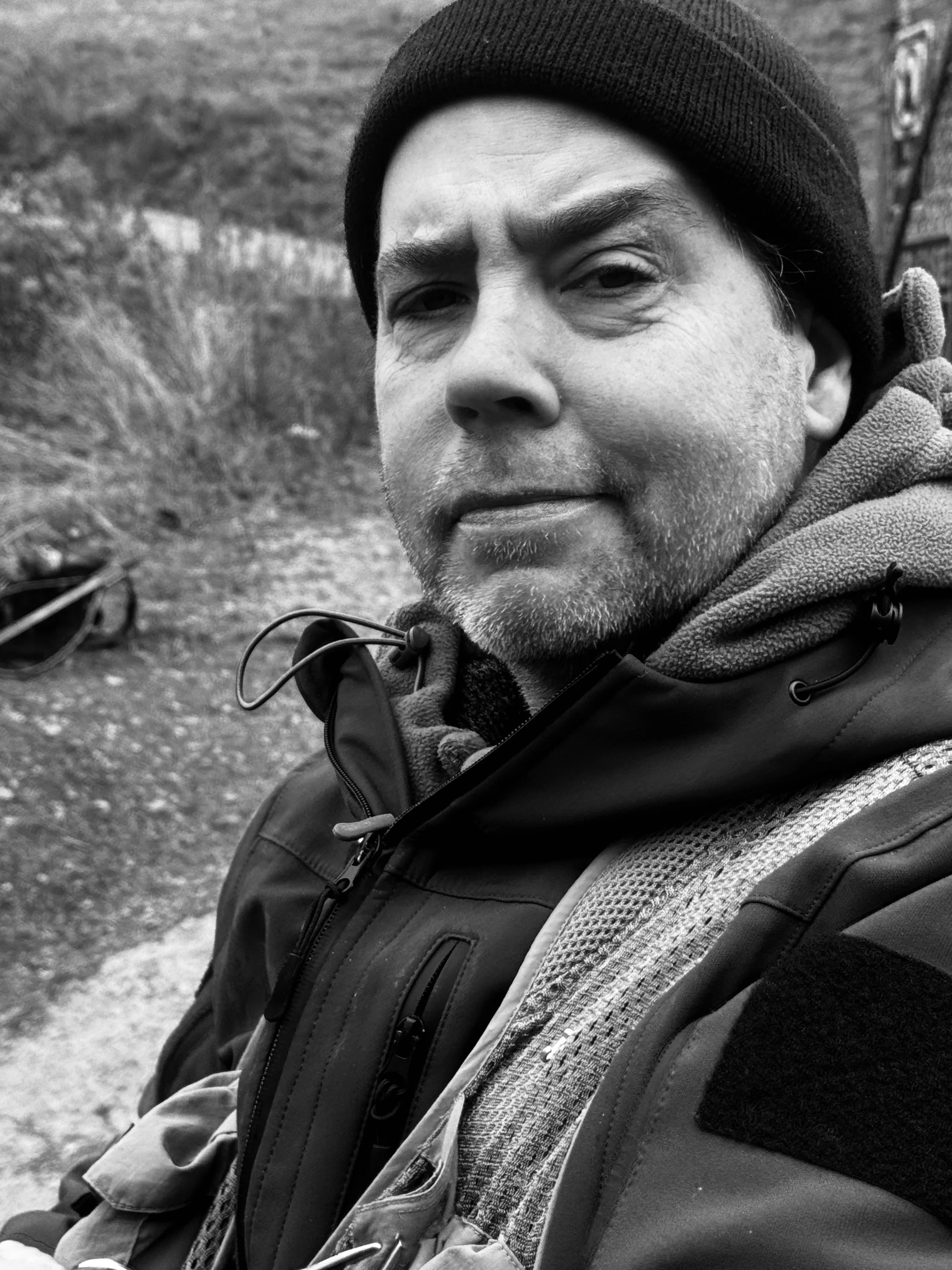 Black and white close-up of a man outdoors wearing a beanie, hoodie, and jacket, with a serious expression, and a blurred natural background.