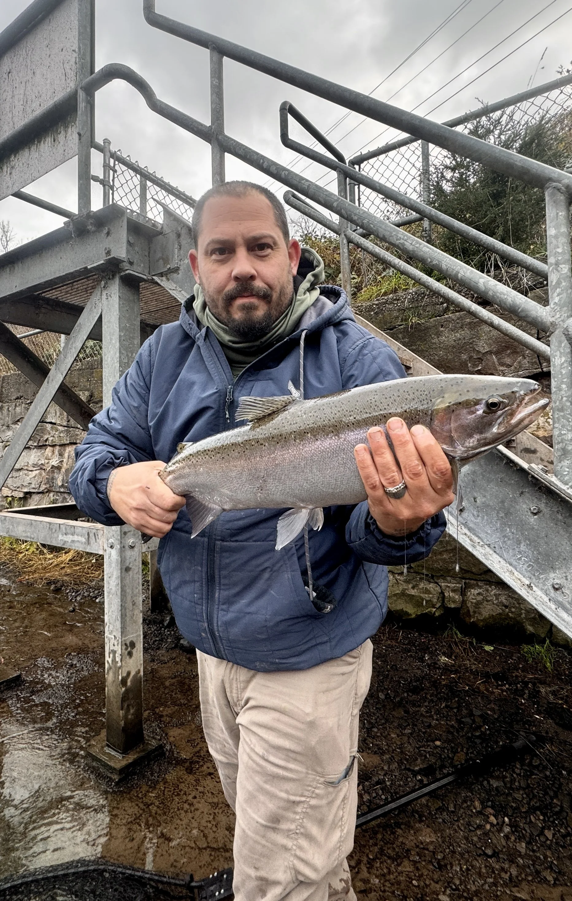 A man holding a large fish, standing outdoors near a metal staircase and a chain-link fence, on overcast day.