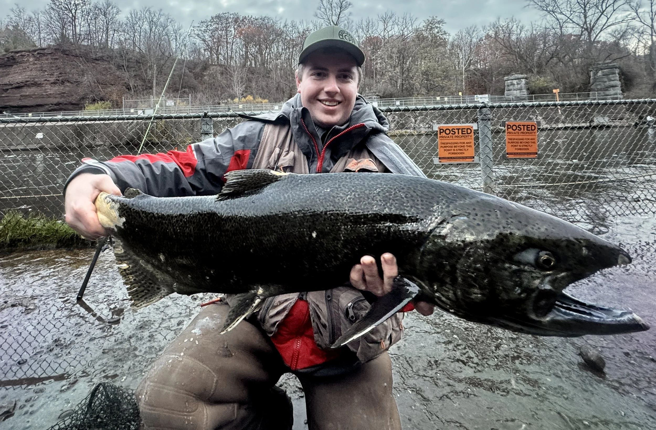 A man smiling and holding a large fish by a river or pond with a chain-link fence and signs in the background.
