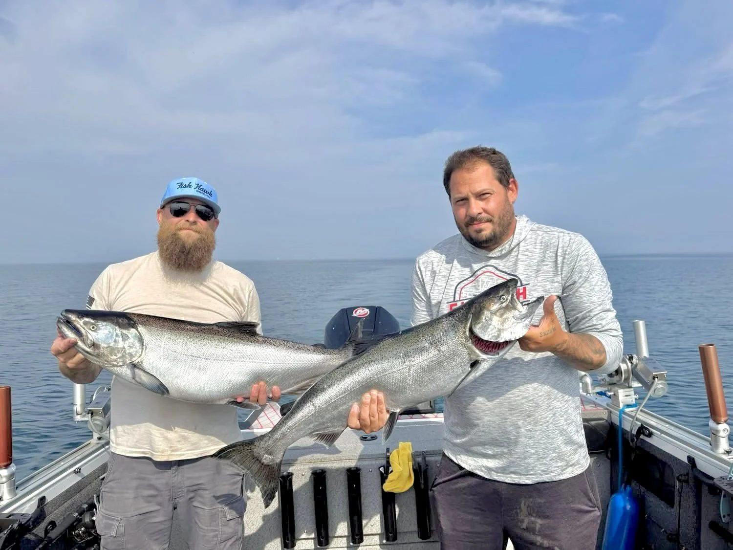 Two men on a boat holding large fish, with a calm ocean and a cloudy sky in the background.