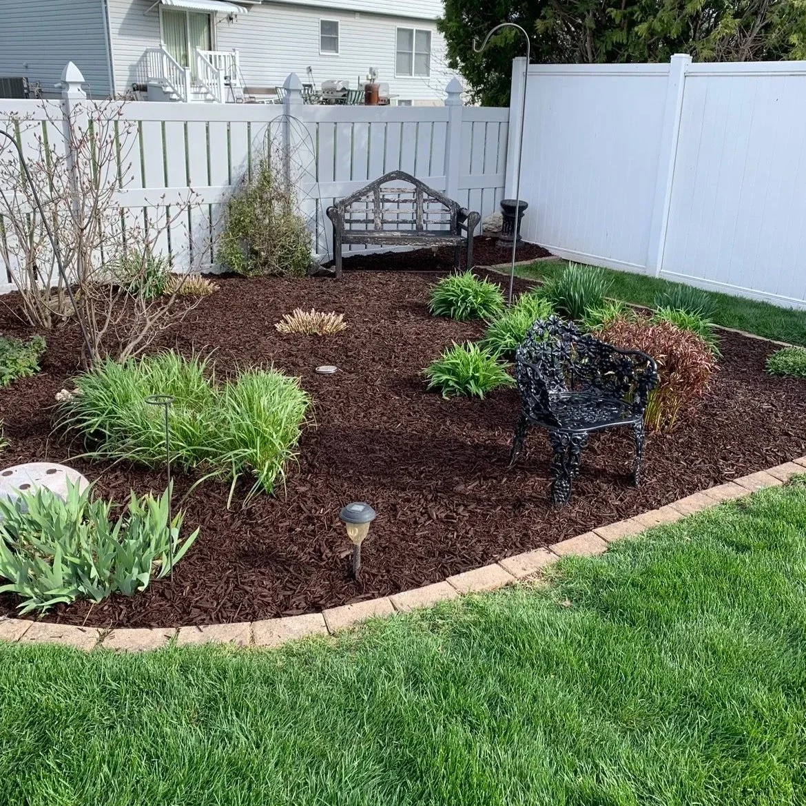 A tidy backyard garden with a white fence, green grass, various plants, a black decorative metal bench, a black garden chair, and a small garden statue.