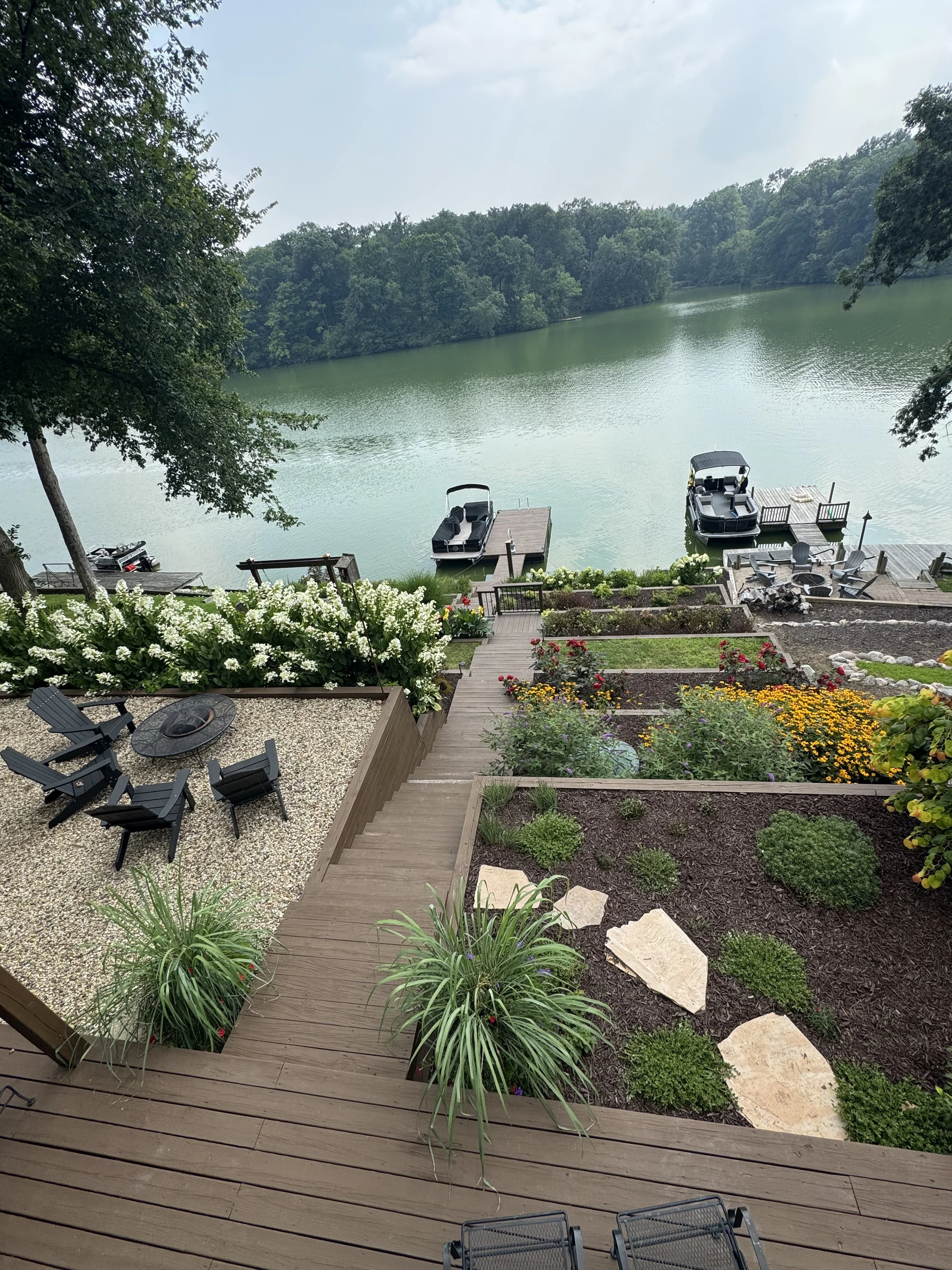 A lakeside backyard with a wooden deck, garden beds with flowers and plants, a gravel area with chairs and a fire pit, and two boats docked at a pier on a calm lake surrounded by trees.