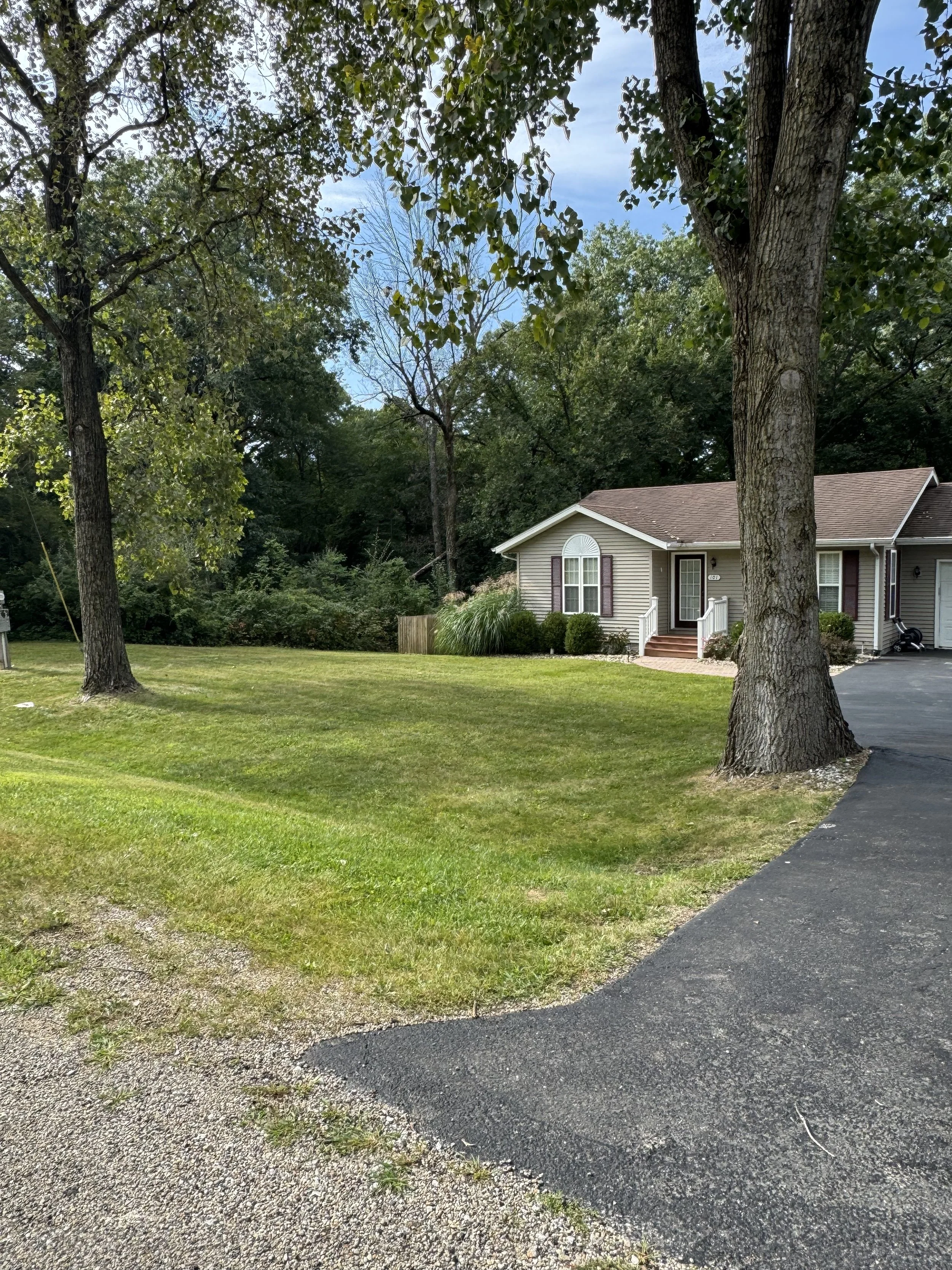 A suburban house with a manicured lawn, trees, and a paved driveway on a partly cloudy day.