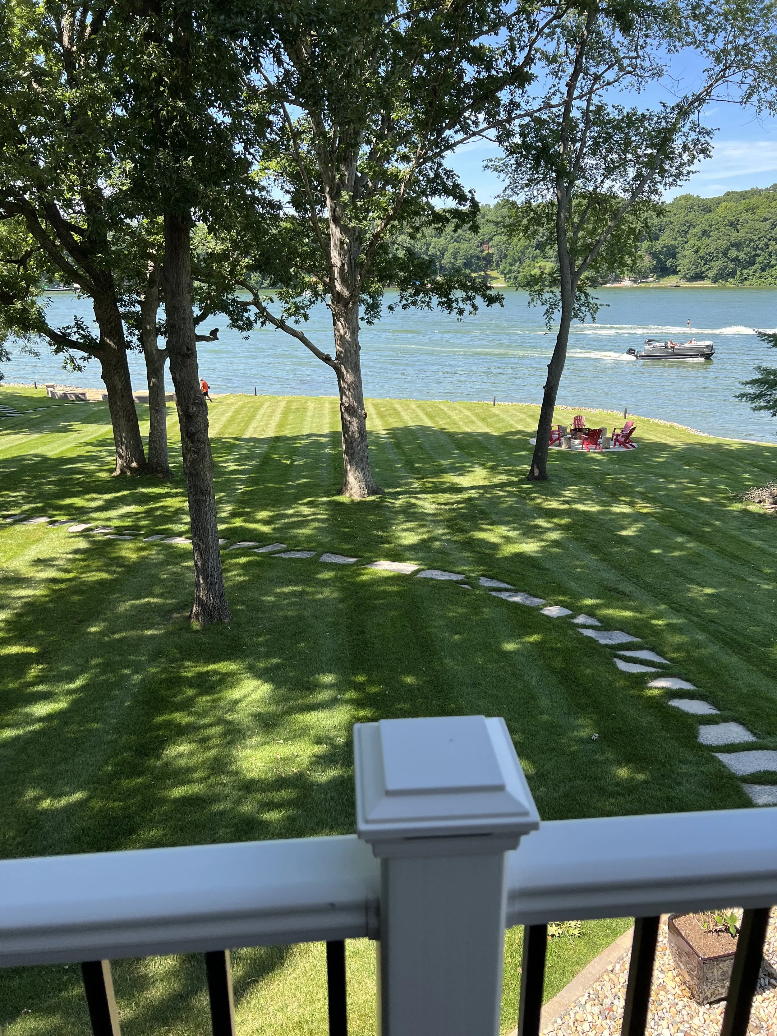 View of a grassy backyard with trees, a paved stepping stone pathway, and red Adirondack chairs in front of a body of water with a boat passing by and a boat wake in the distance, seen from a balcony with a white railing.