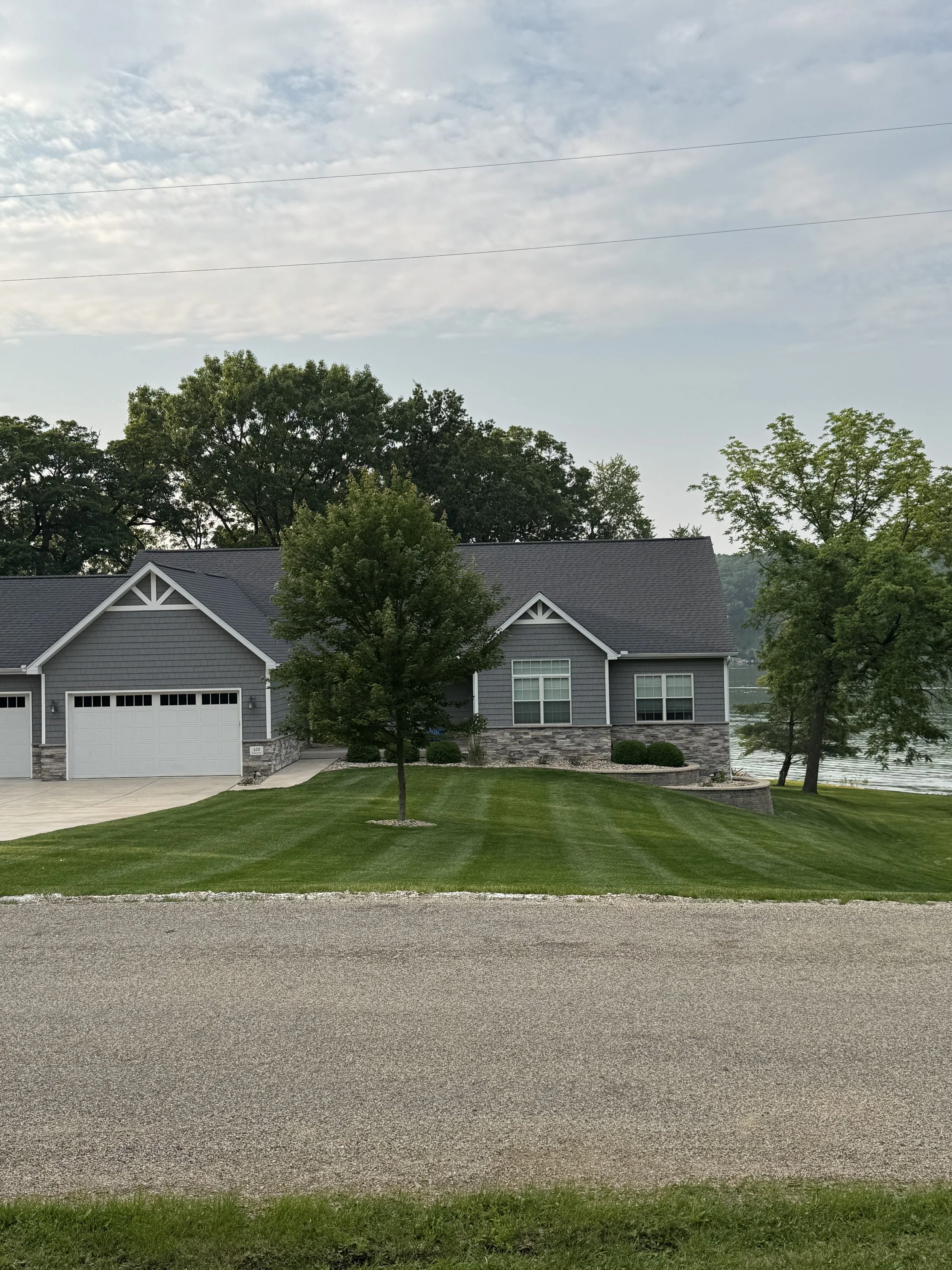 A two-story gray house with a garage, surrounded by green grass, trees, and a river in the background under a cloudy sky.