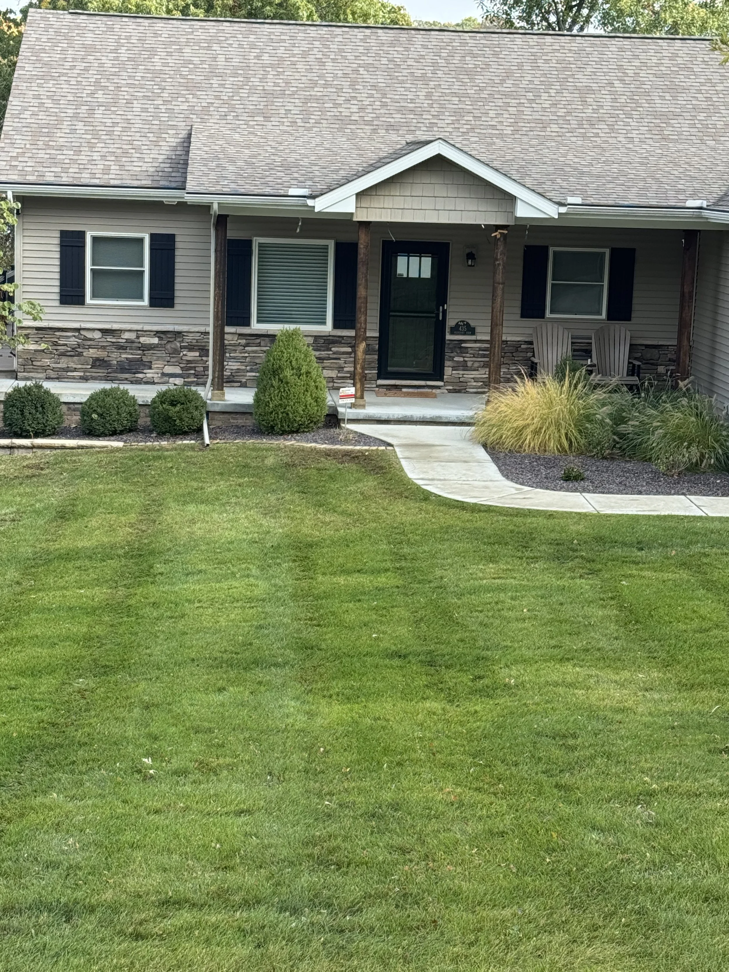 Front view of a house with a stone and siding exterior, a small covered porch with two chairs, a curved concrete walkway, a well-maintained lawn, and landscaped bushes and plants.
