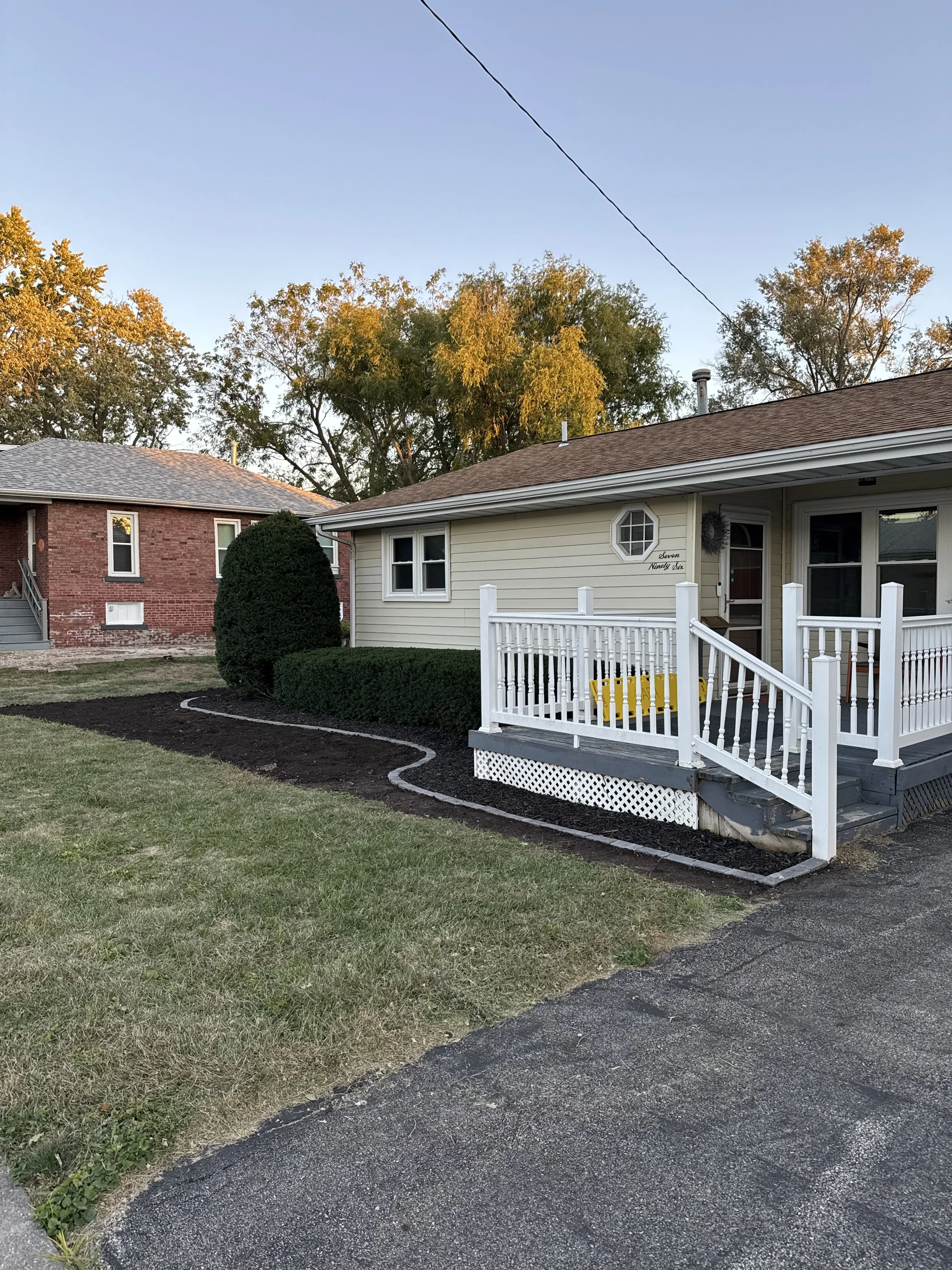 A small residential house with a beige exterior, white railing porch, and a yellow swing. The yard has freshly turned soil with a garden border and trimmed bushes, and a neighboring brick house is visible. Trees with autumn leaves are in the backgrou