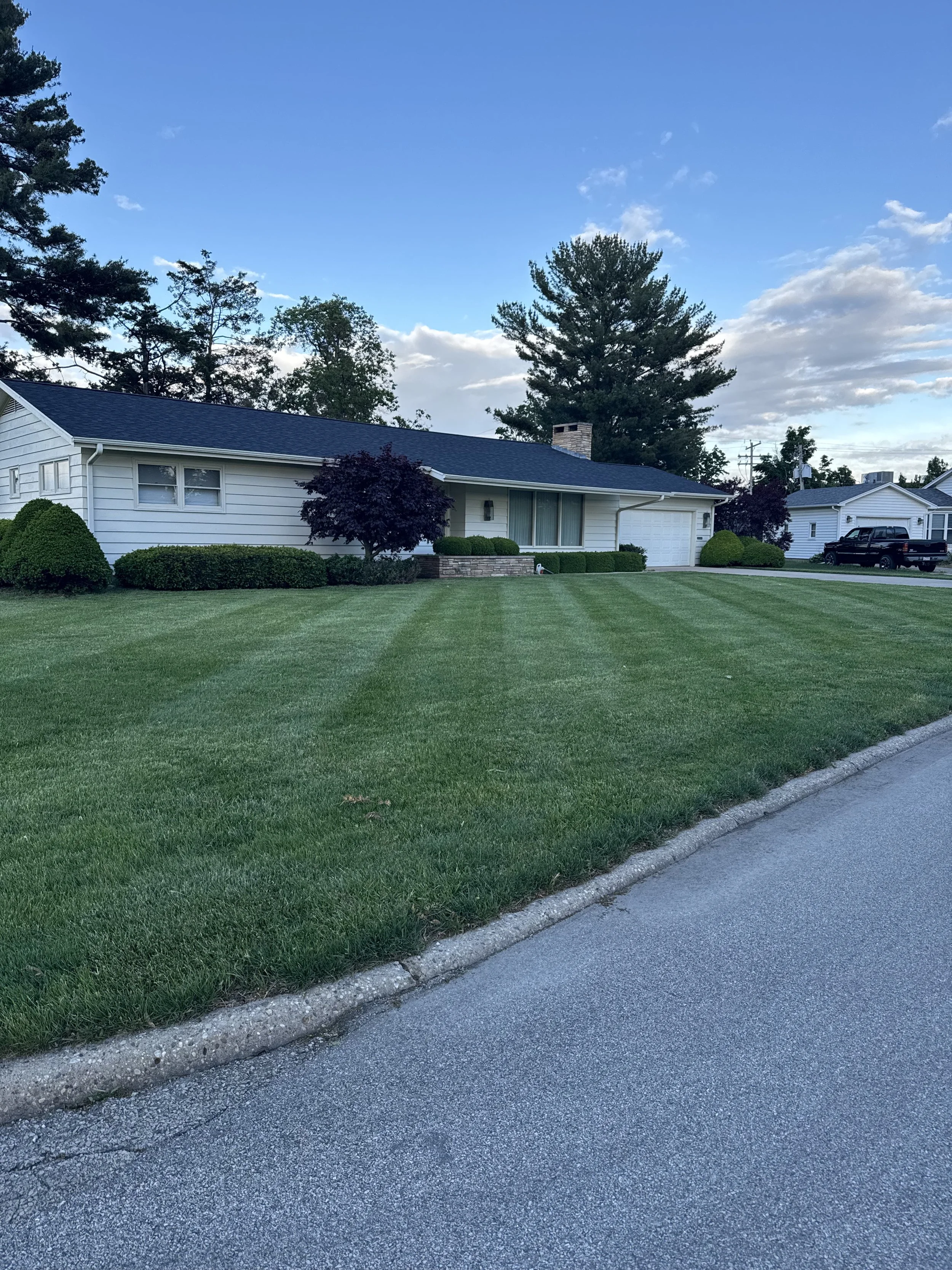 A suburban house with a white exterior, dark roof, well-manicured lawn, bushes, and trees, with a driveway on the right and a clear blue sky above.