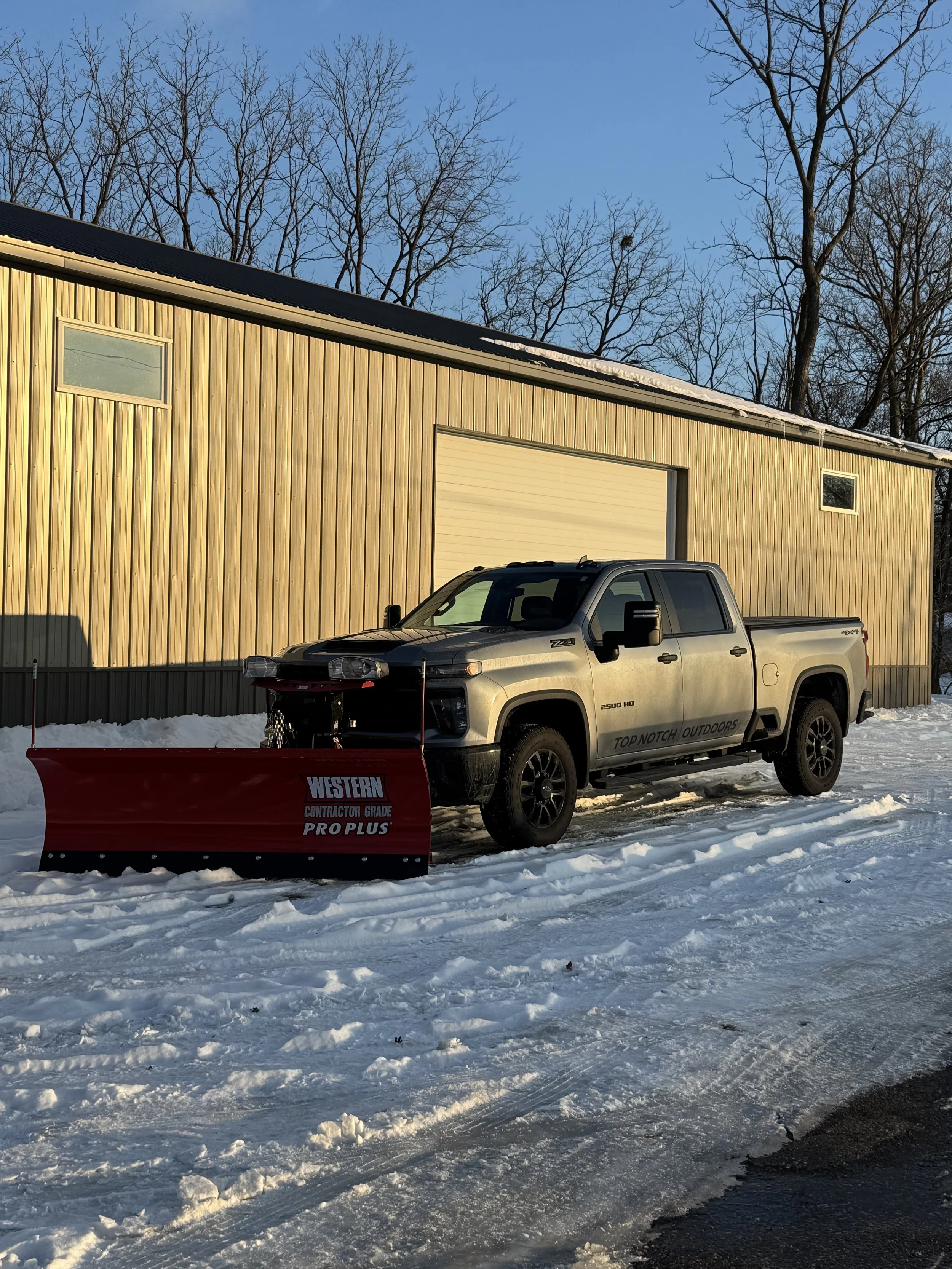 A white pickup truck with black wheels and side mirrors parked on snow in front of a yellow wooden building. The truck has a red snow plow attached to the front labeled 'Western Contractor Grade Pro Plus.' The background features leafless trees and a