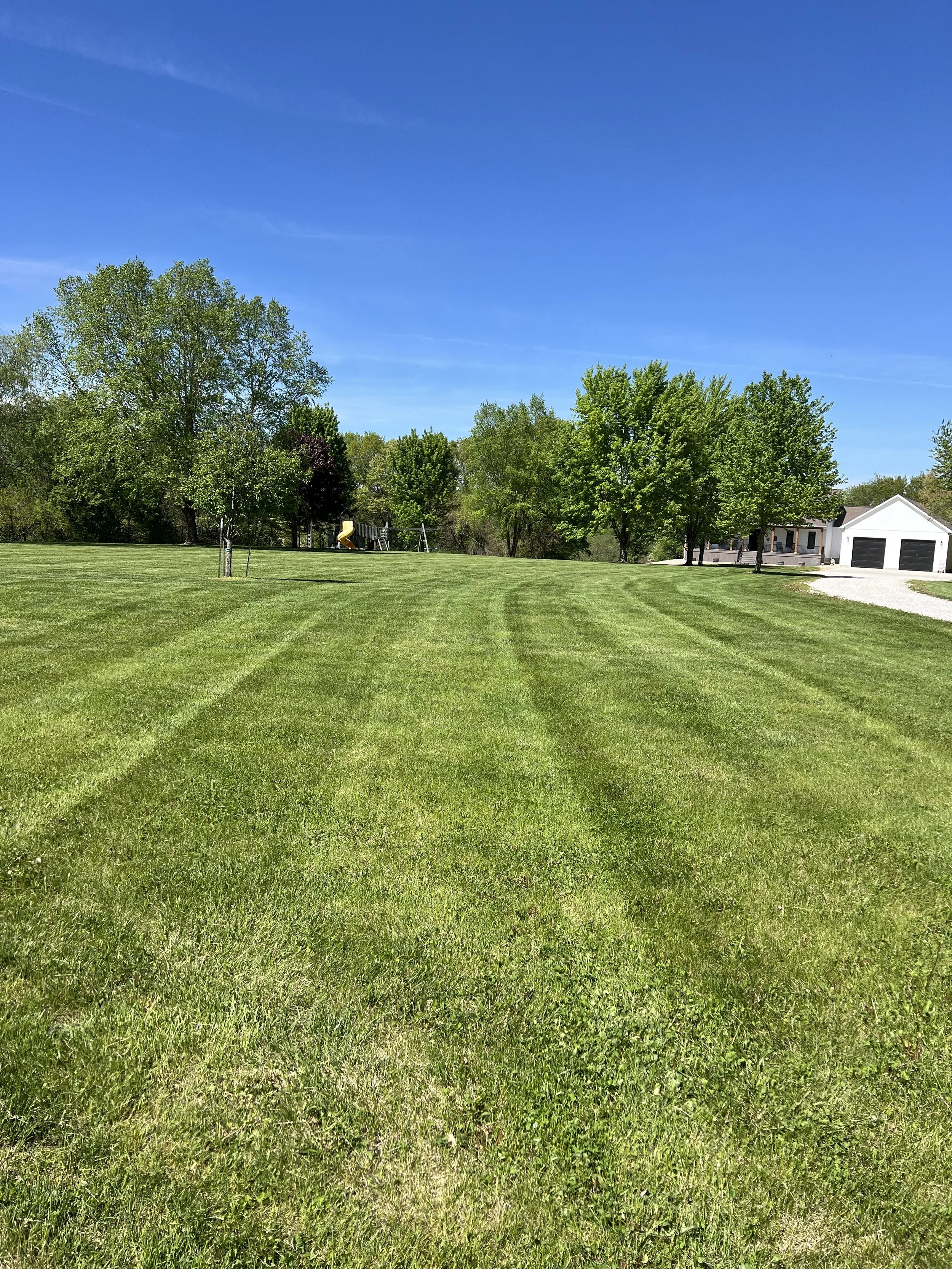 A wide, freshly mowed grassy lawn with visible mower lines extends toward a line of deciduous trees and a white house with black garage doors under a clear blue sky.