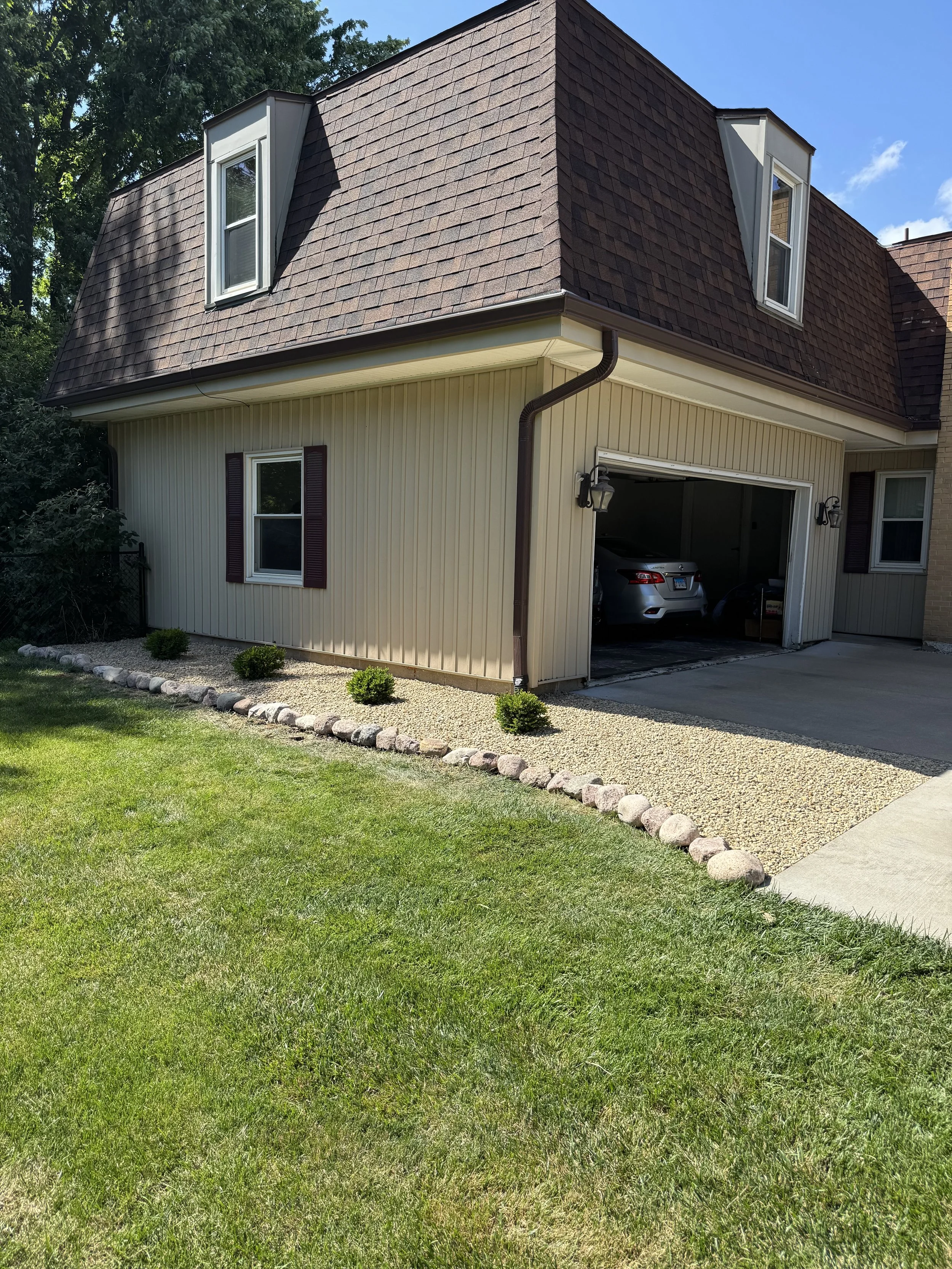 Front view of a house with beige siding, brown roof, and a garage with a car inside, surrounded by a lawn and landscaped with rocks and small bushes.