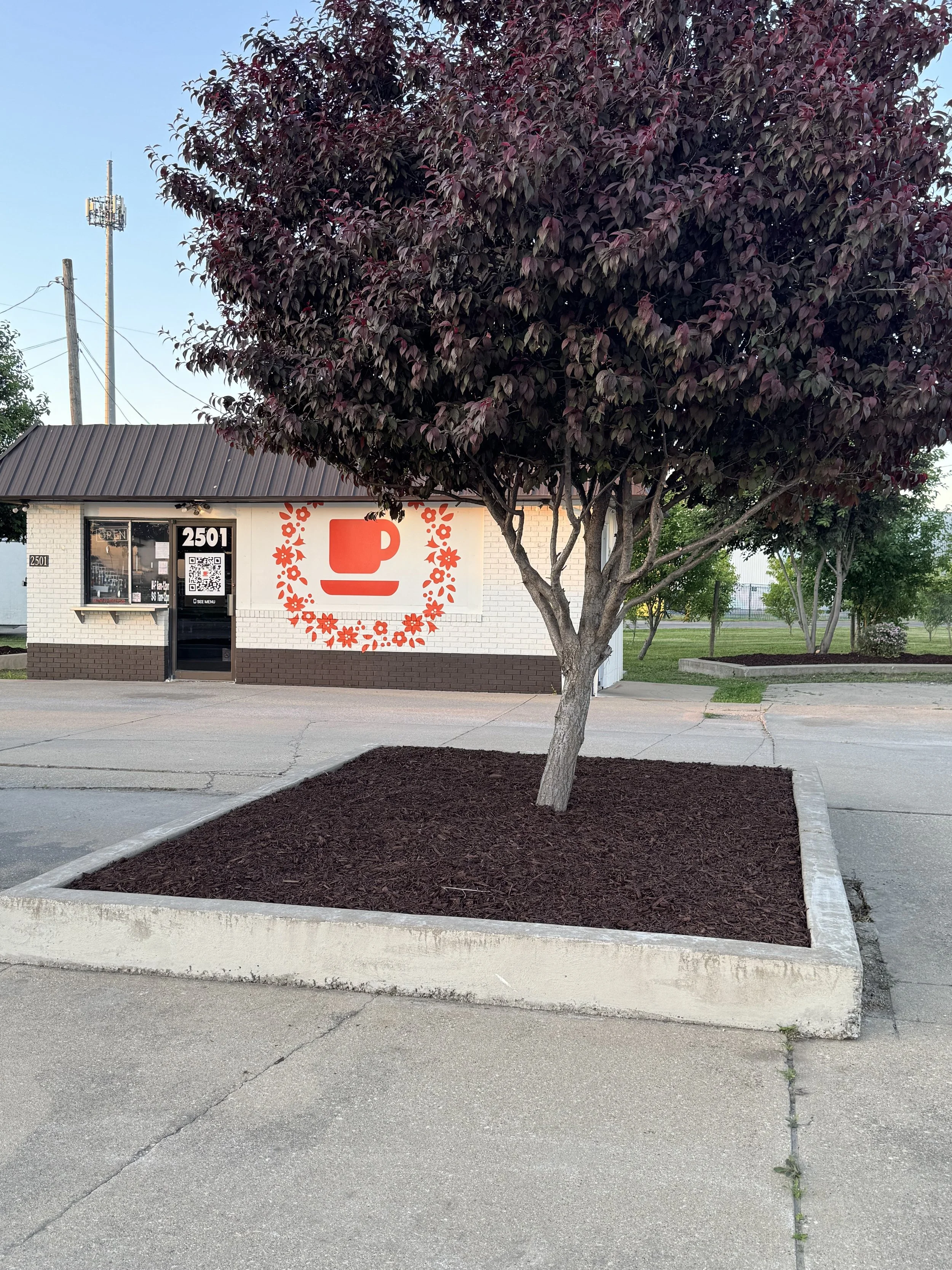 A small white building with a black roof, a large red logo of a coffee cup with a heart-shaped steam, and a QR code on the front window. There is a pink-leaved tree in a rectangular planter in front and a parking lot.