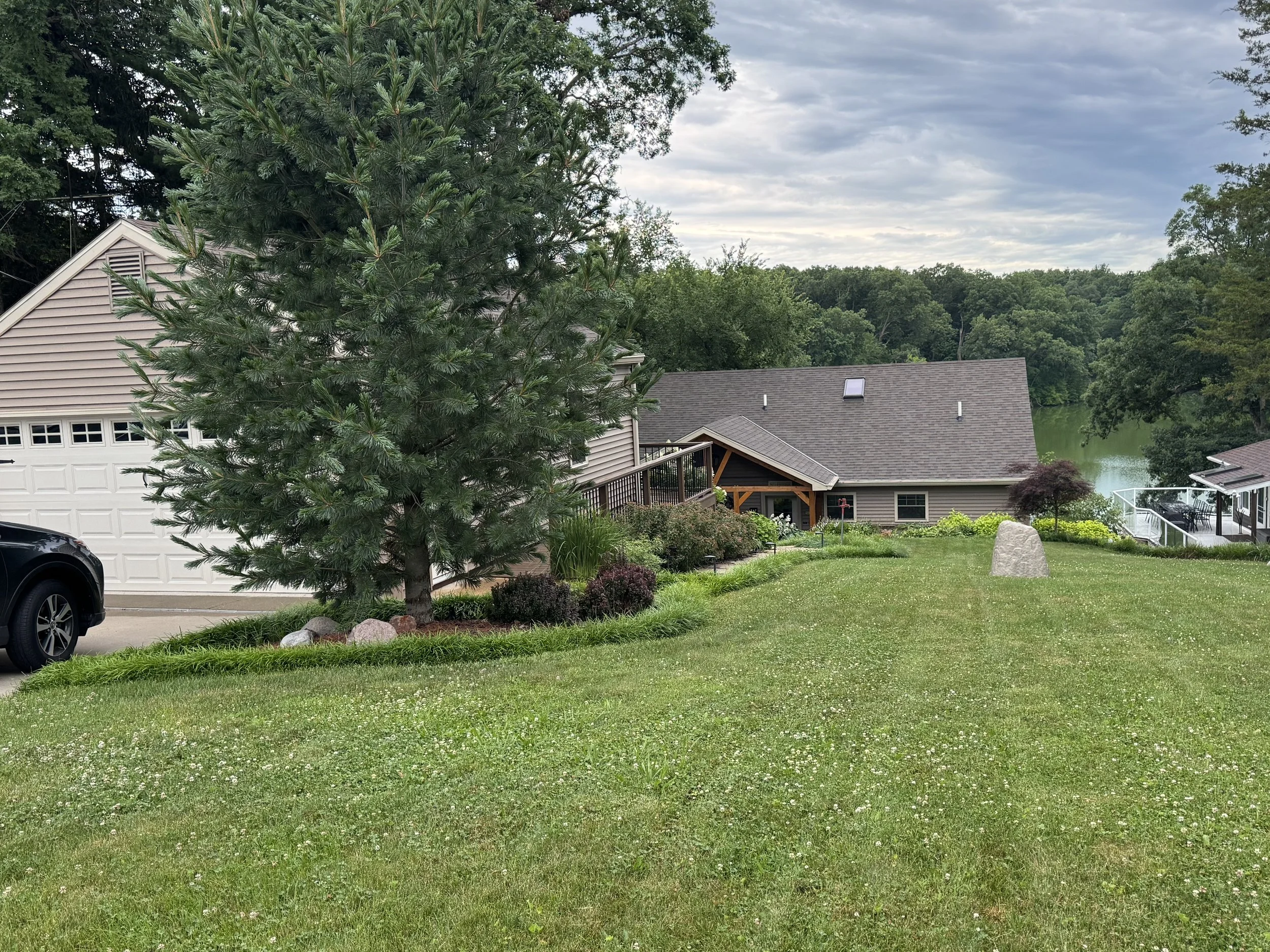 A backyard scene with a large pine tree, a manicured lawn, a stone monument, and a house with a deck overlooking a body of water, surrounded by trees under a cloudy sky.