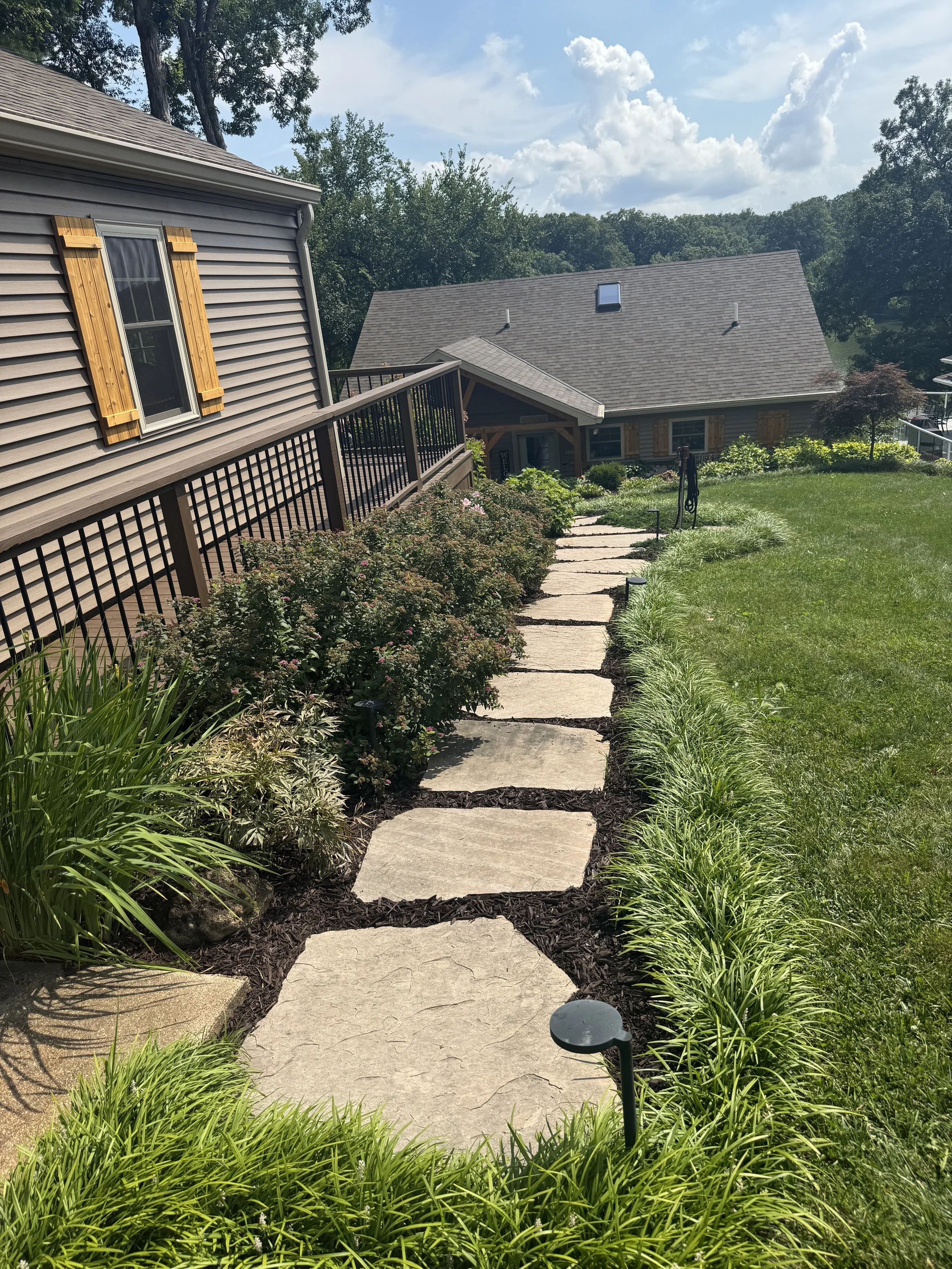 A stone pathway with grass and garden plants on either side leads downhill past a house with brown shutters and a landscaped yard under a partly cloudy sky.