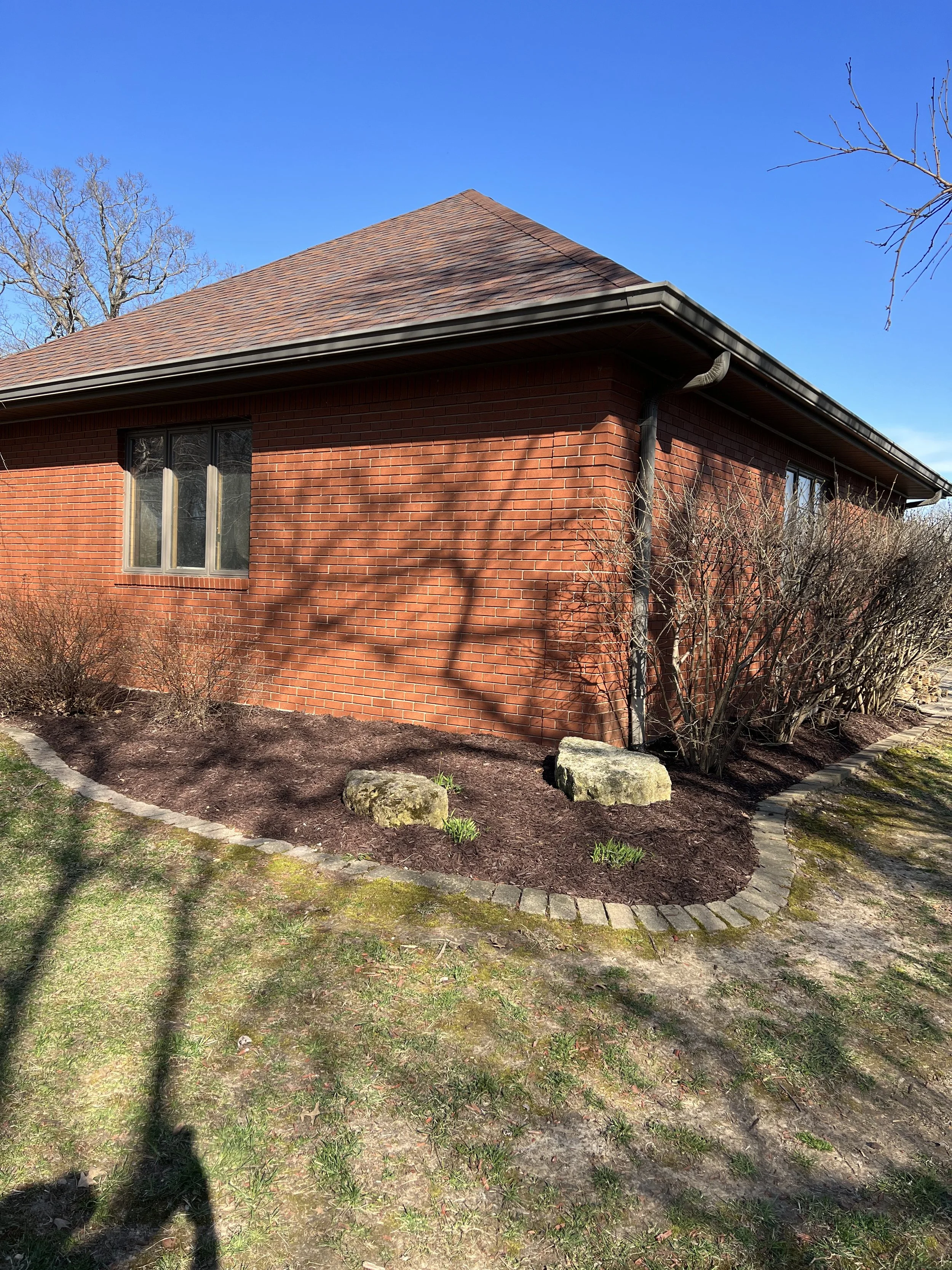 Side of a brick house with a brown shingle roof, two windows, a small garden with shrubs and rocks, and a partly visible sidewalk.