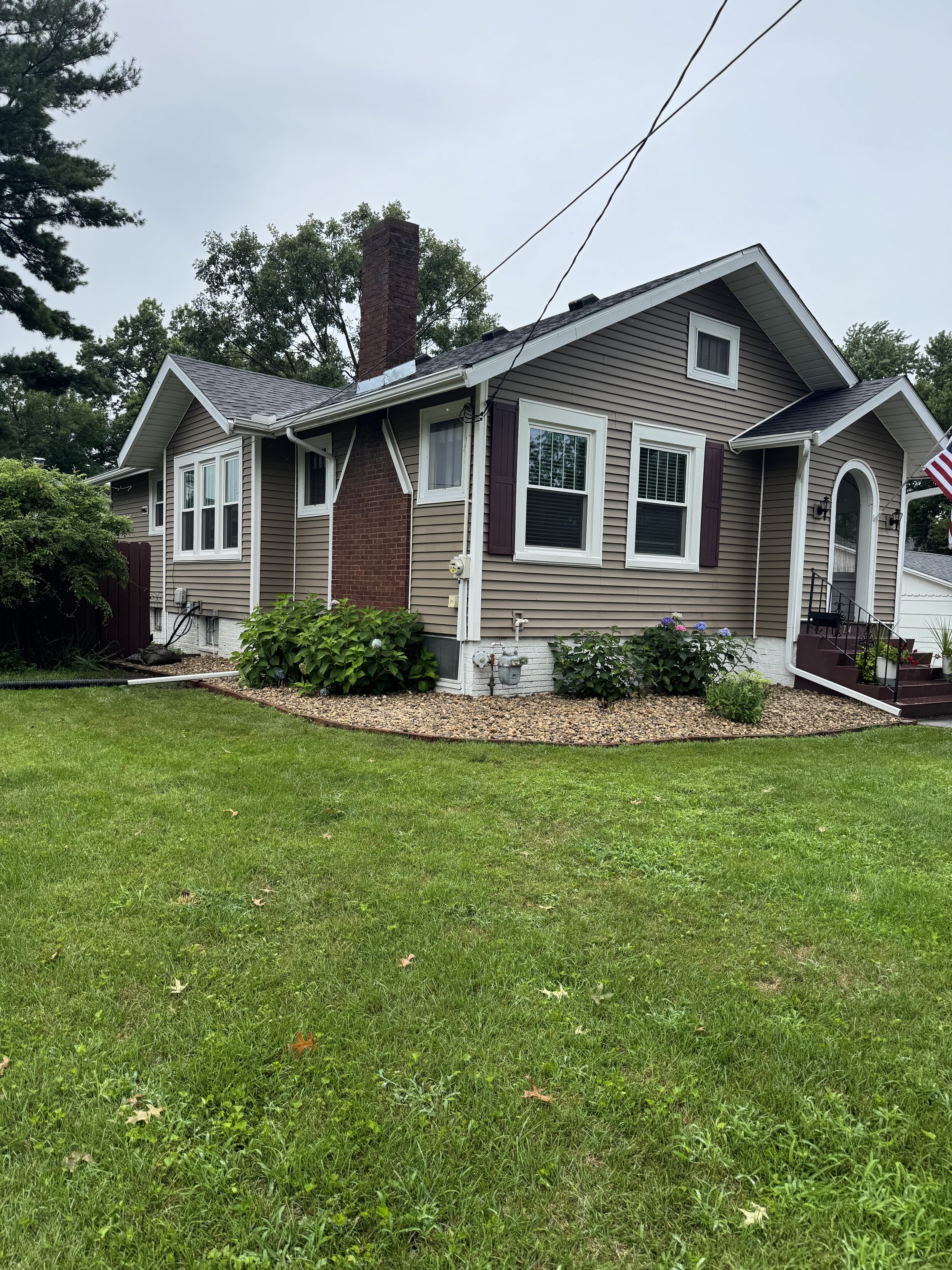 A single-family house with beige siding, purple shutters, and a brick chimney, surrounded by a green lawn and landscaped garden beds with bushes and flowers.