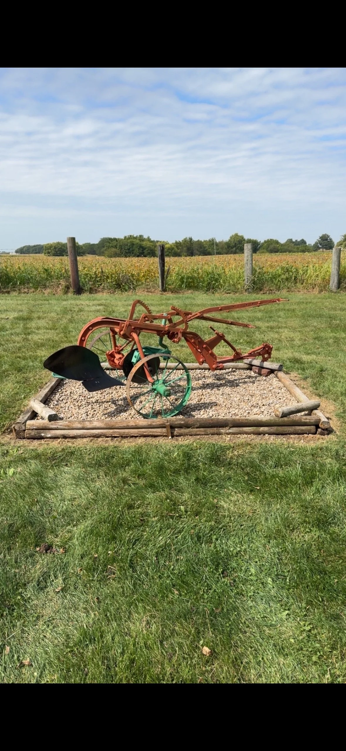 An old, rusted agricultural machine, possibly a plow or cultivator, displayed on a small gravel patch, outdoors on grass with a field and blue sky in the background.