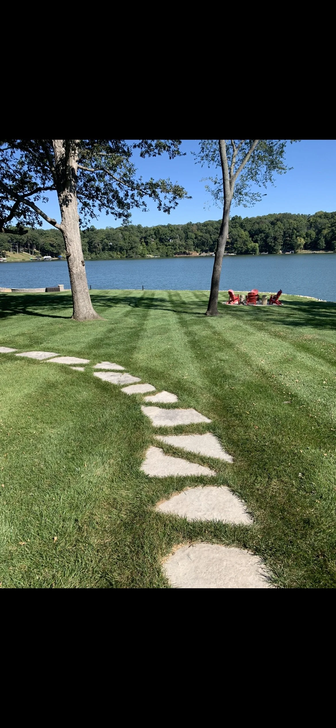 A lakeside scene with green grass, a stone pathway, two trees, a lake, and a red picnic table with chairs.