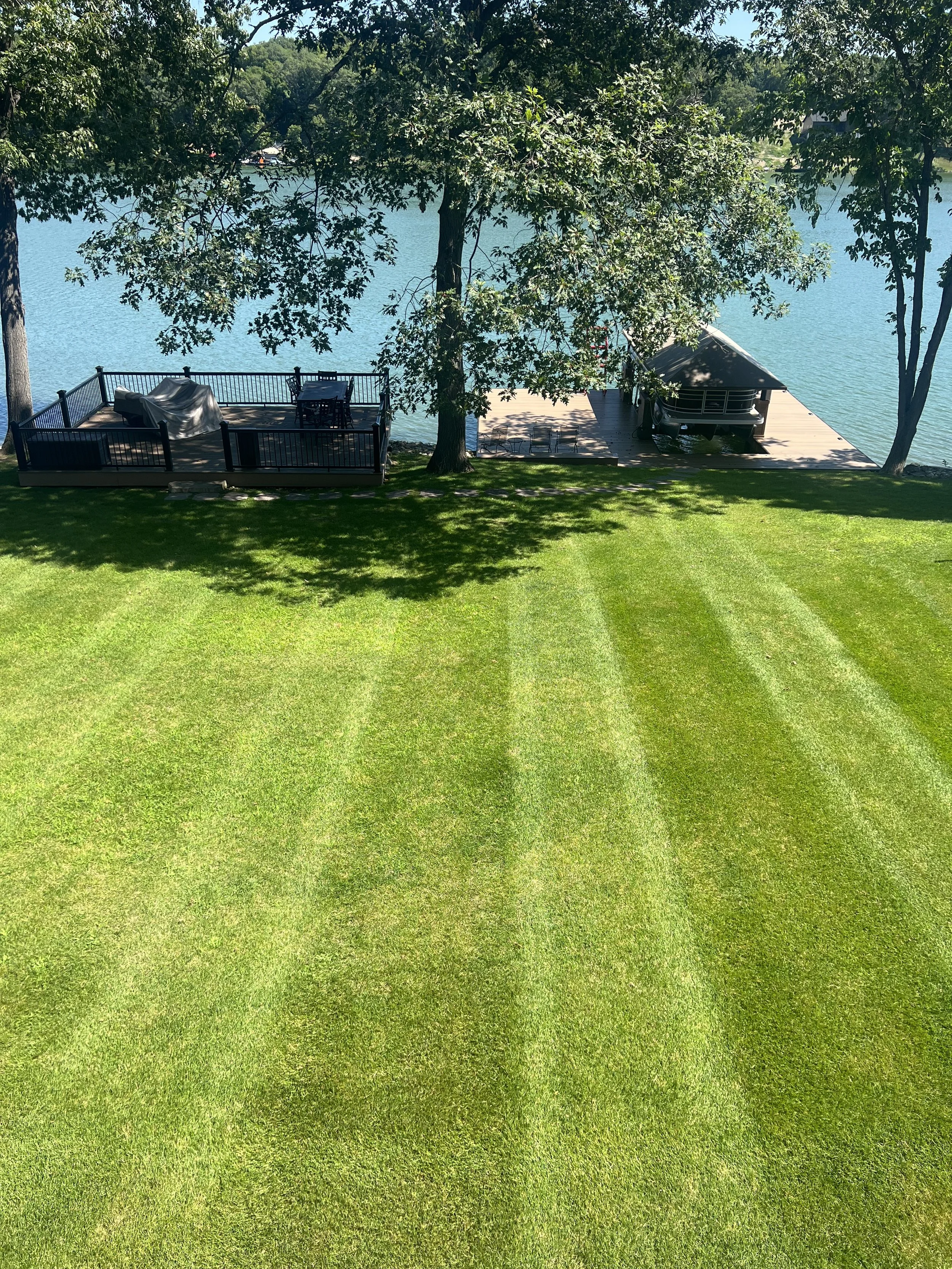 Lush green backyard with striped grass, overlooking a calm lake. There is a wooden deck with a covered grill, a patio table, and chairs. A small dock extends onto the lake, with a boat docked underneath a gazebo.