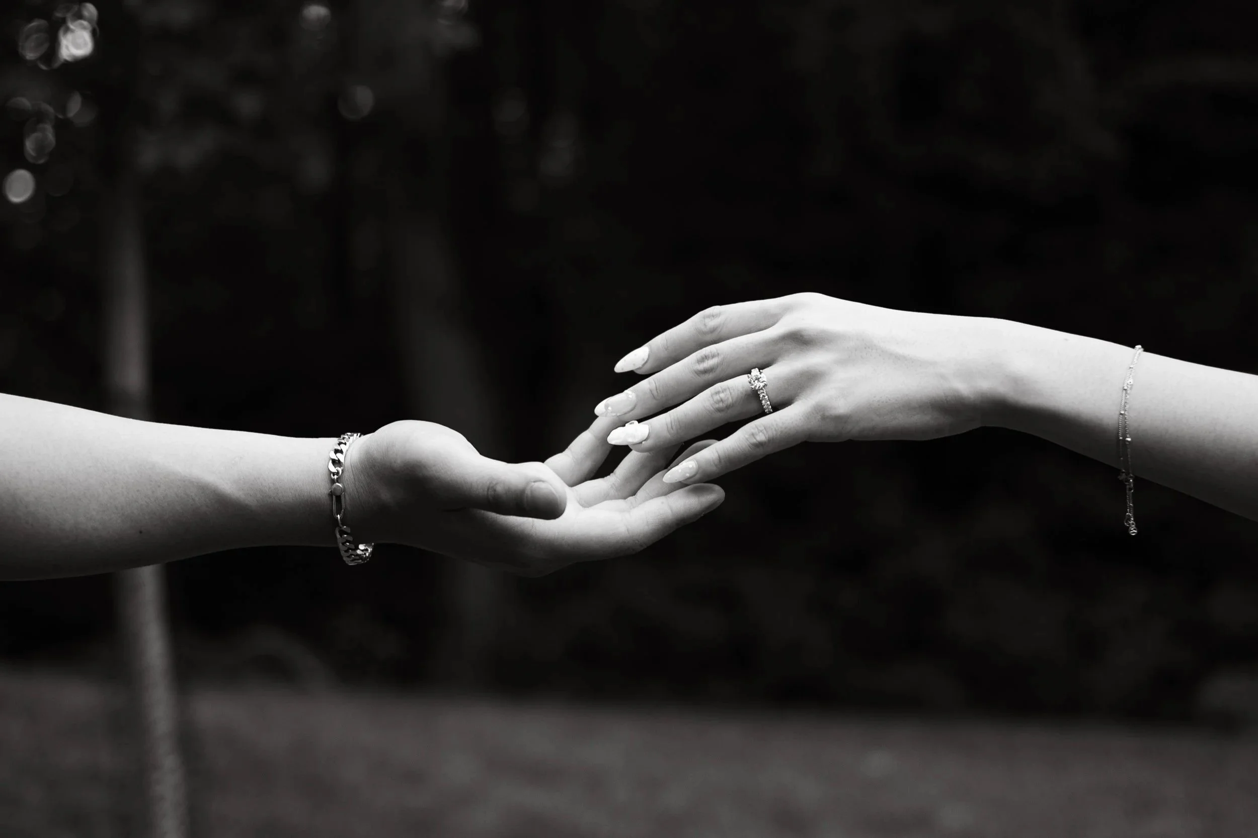 Black and white photo of two hands reaching out to touch each other, adorned with jewelry, against a dark blurred background.