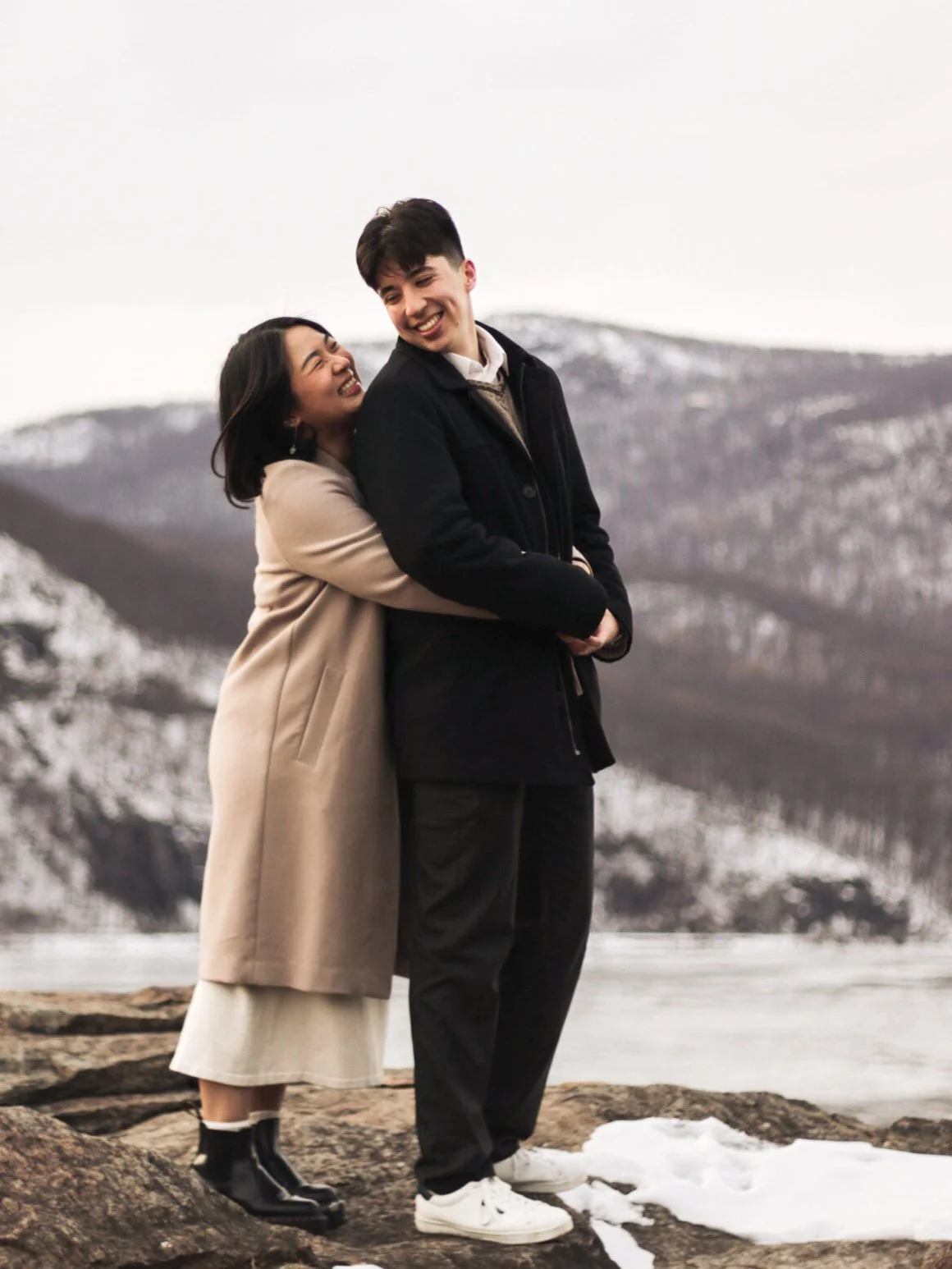 A couple standing outdoors on a rocky shore with snow patches, embrace and laugh, with snow-capped mountains in the background during winter.