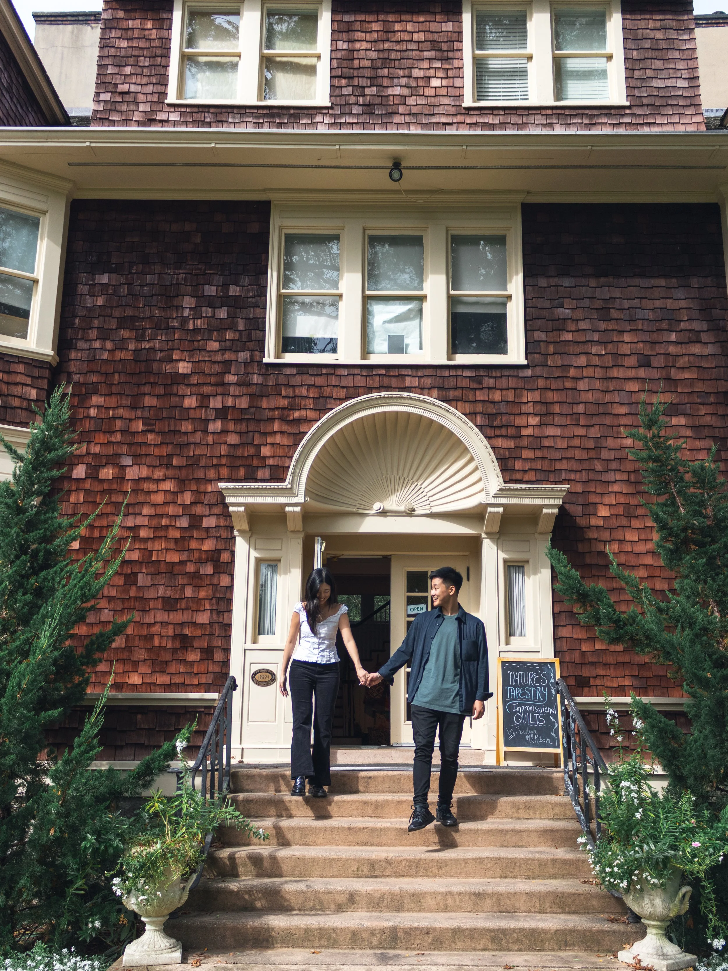 A man and woman holding hands on the front steps of a red brick house with white trim and large windows, surrounded by greenery and flowers.