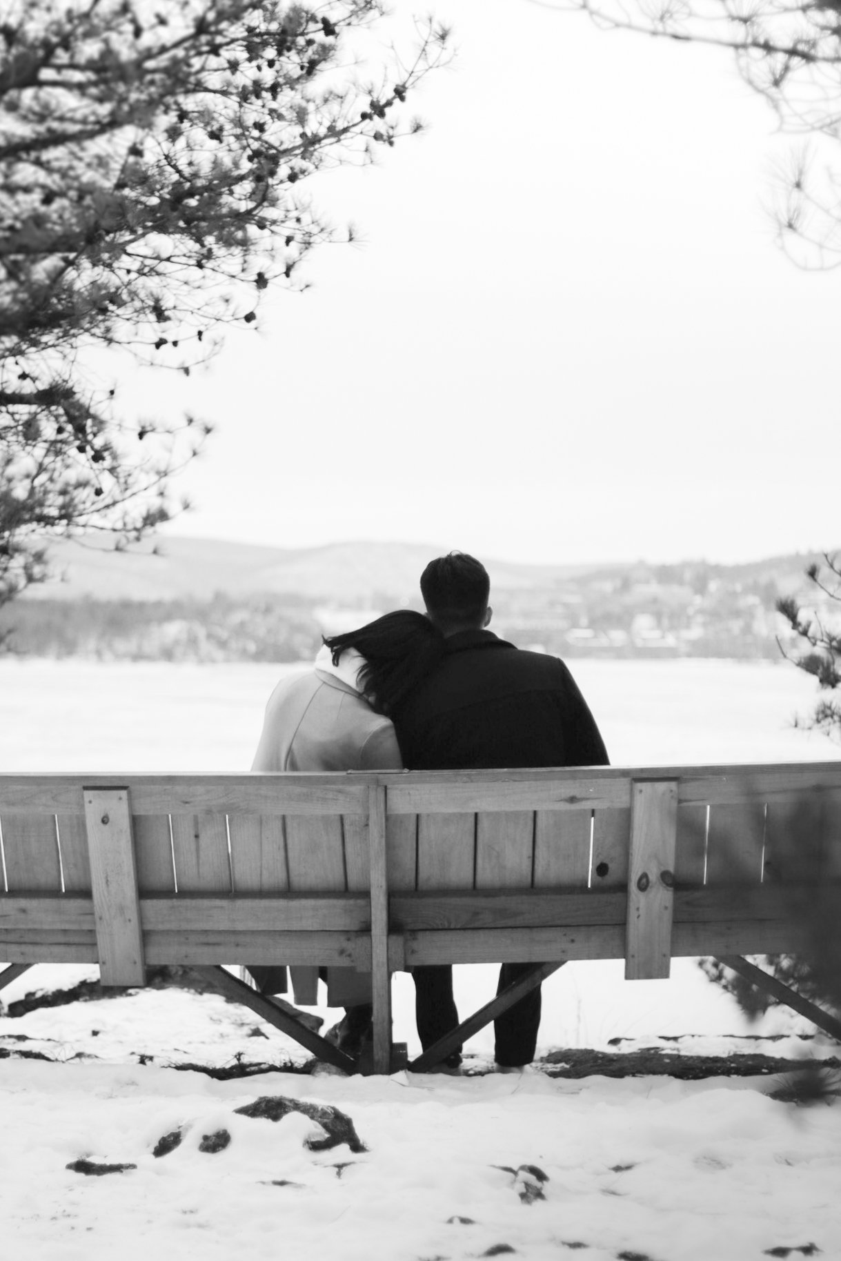 A black and white photo of a couple sitting on a wooden bench outdoors in a snowy landscape, with trees overhead and mountains in the distance, embracing and looking at a scenic view.