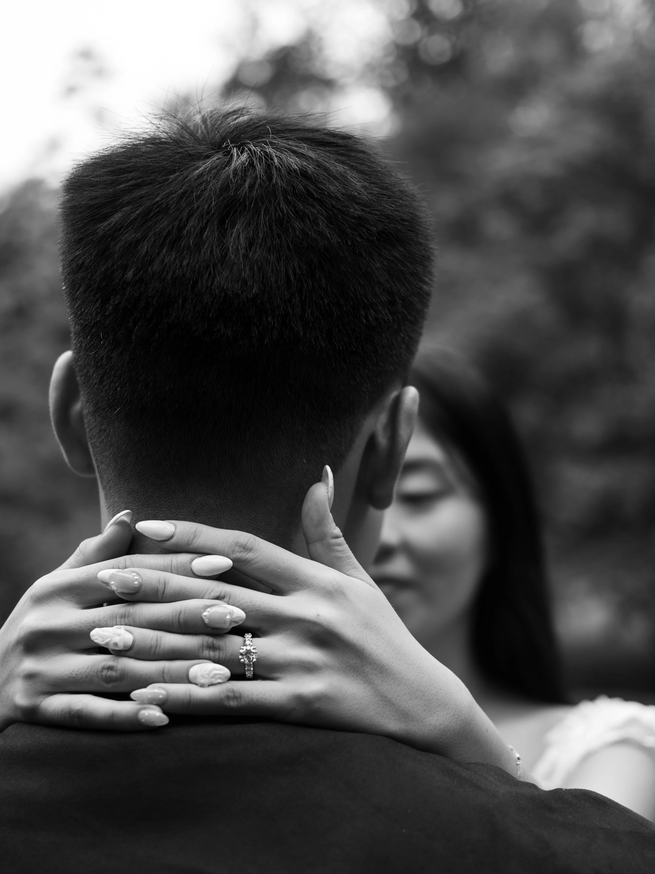 A close-up black-and-white photo of a person with short hair embracing another person, with only the back of the head visible. A woman's hand with a wedding ring gently rests on the man's shoulder.