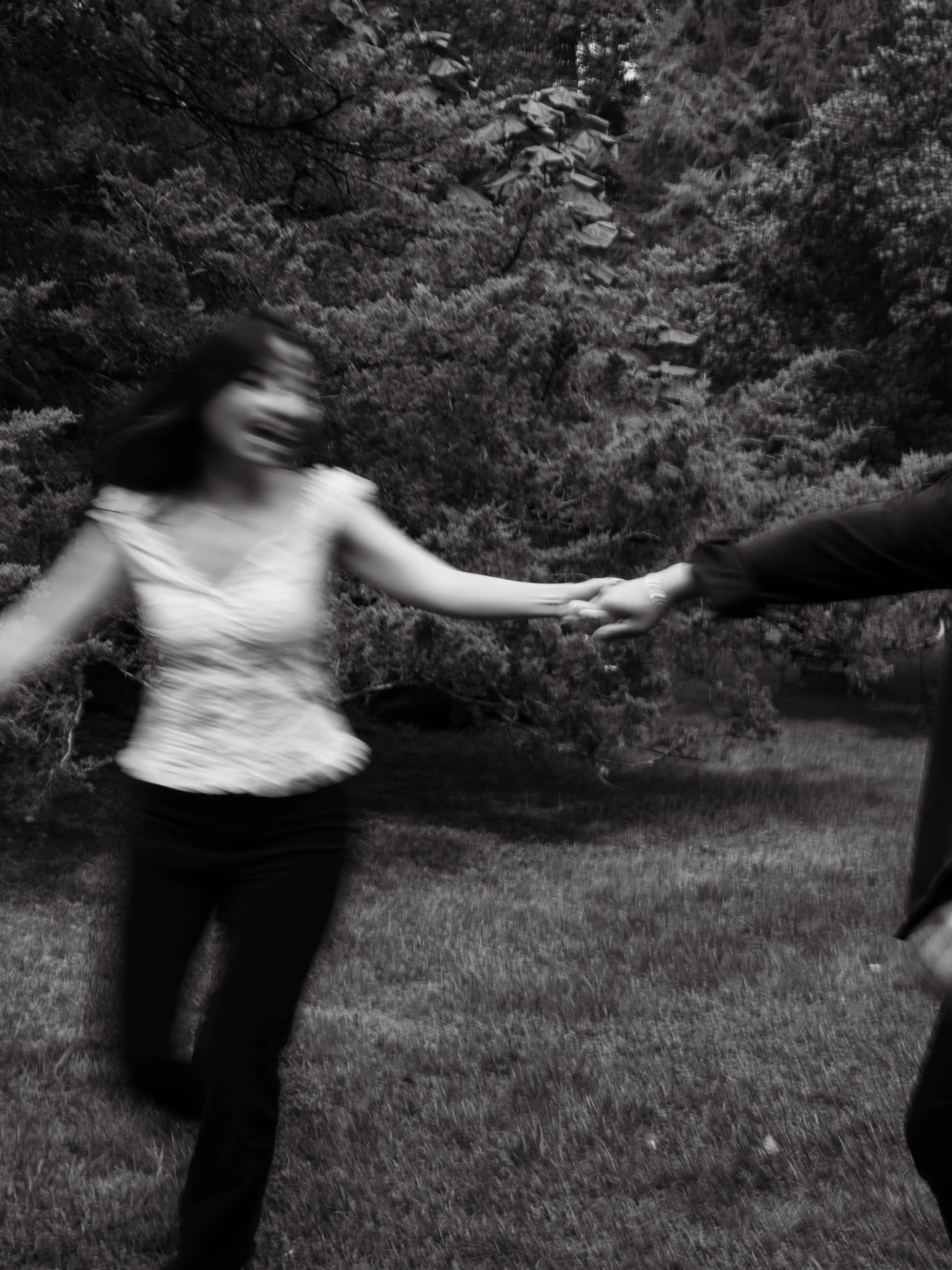 A blurred black and white photo of a woman in a white dress holding hands with a man in a dark suit, outdoors with trees and grass.