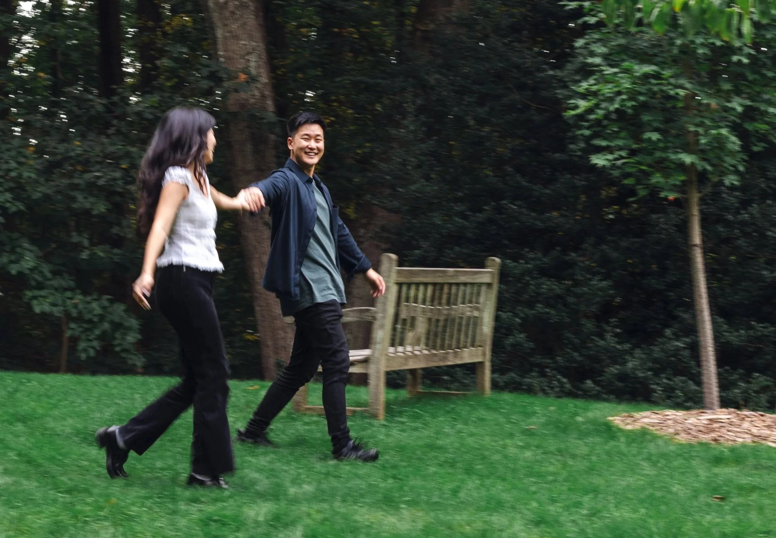 Two people, a man and a woman, holding hands and running through a park with green grass and trees.