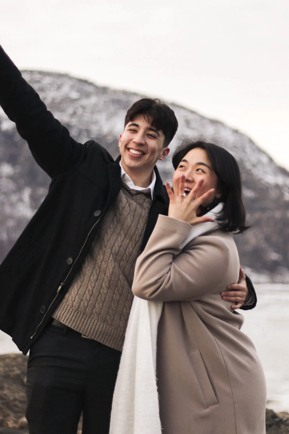 A smiling young man and woman standing outdoors, taking a selfie together in front of snow-covered mountains.