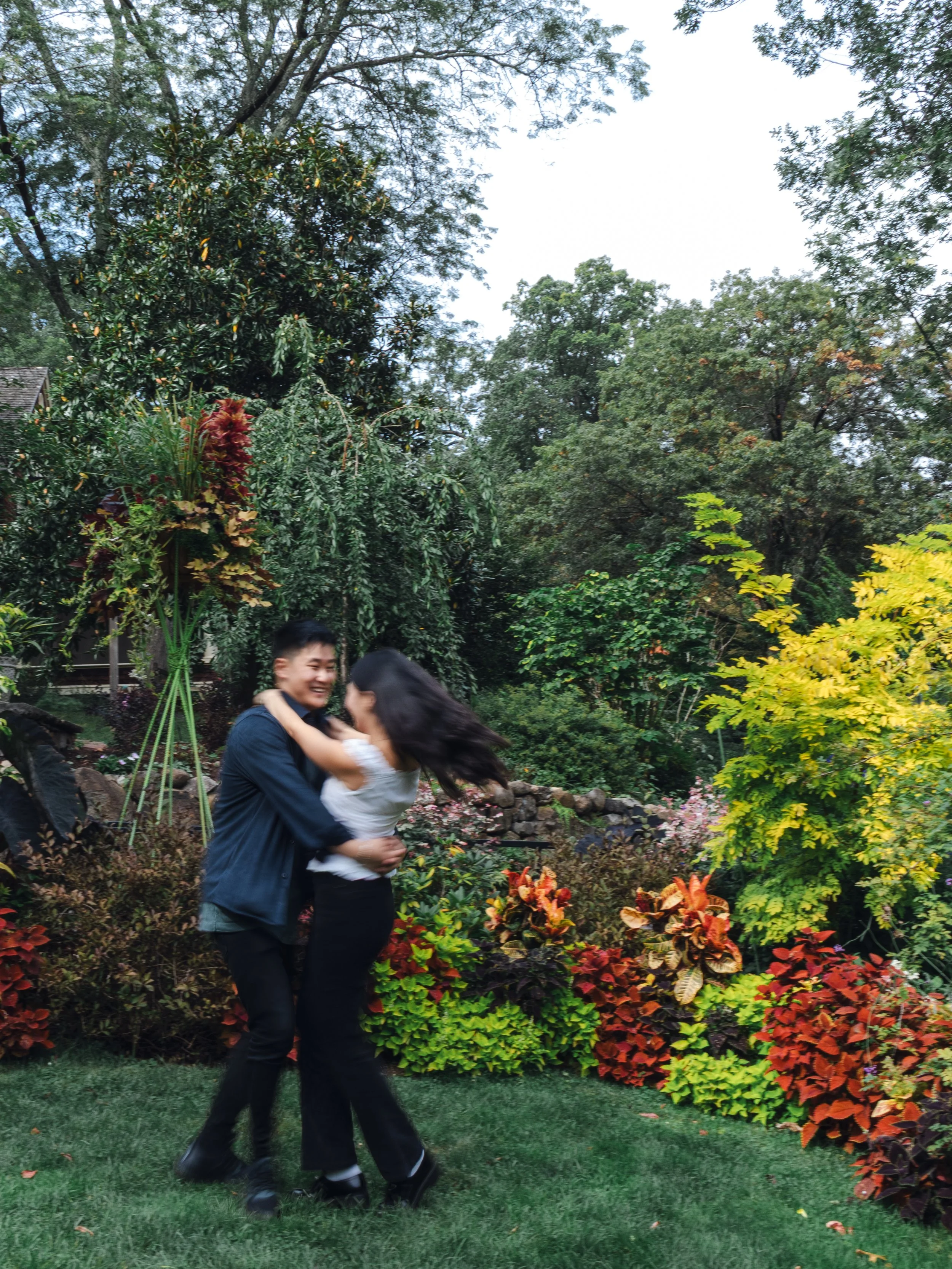 A young man and woman are dancing outdoors in a garden with colorful plants and trees, smiling and enjoying each other's company.
