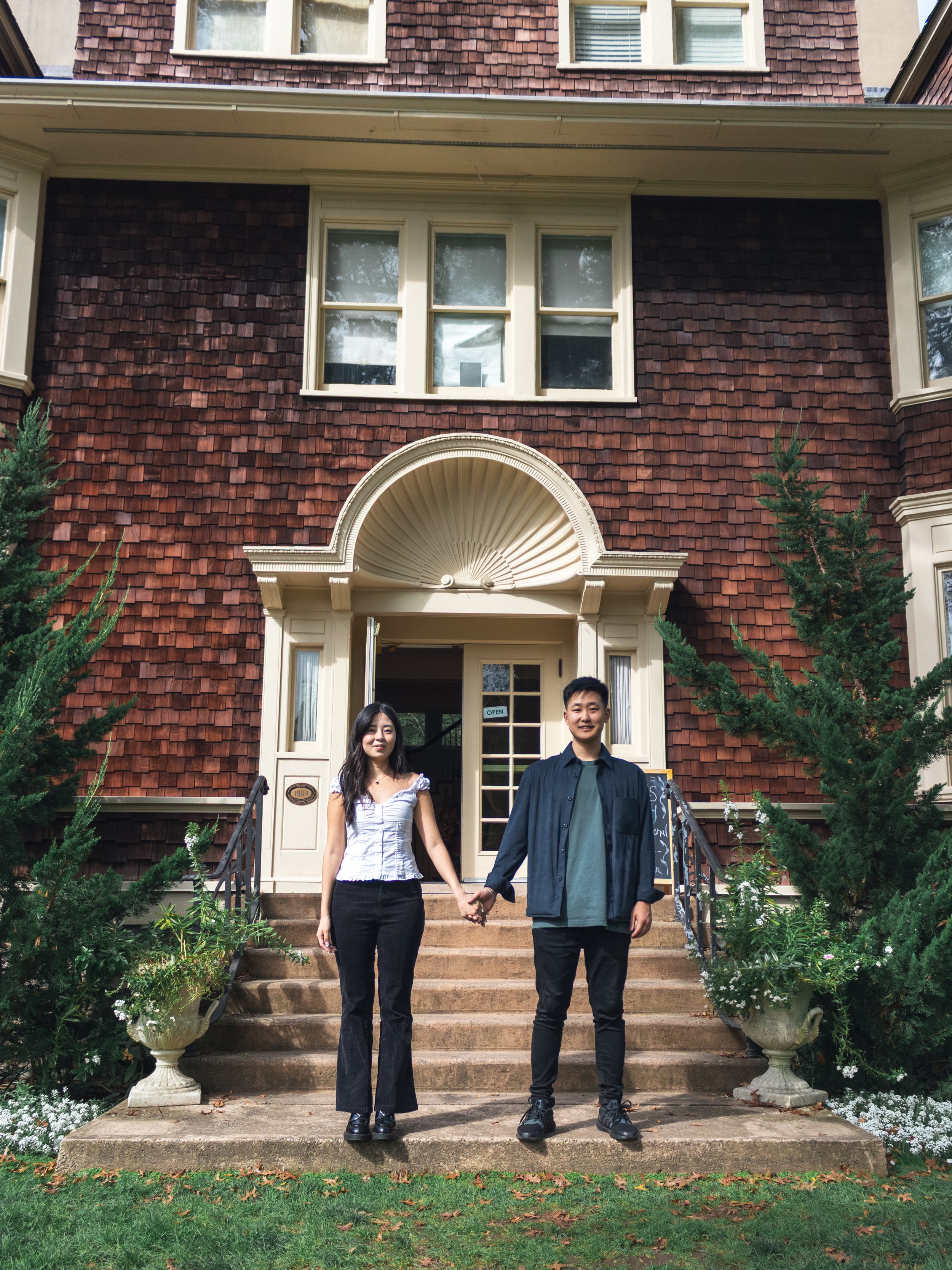 A young woman and a young man holding hands standing on the steps of a house with a brown shingled exterior, white trim, and large potted plants on either side of the stairs. The woman is wearing a white blouse and black pants, and the man is wearing a black jacket and pants. The house has an arched decorative detail over the door and multiple windows.