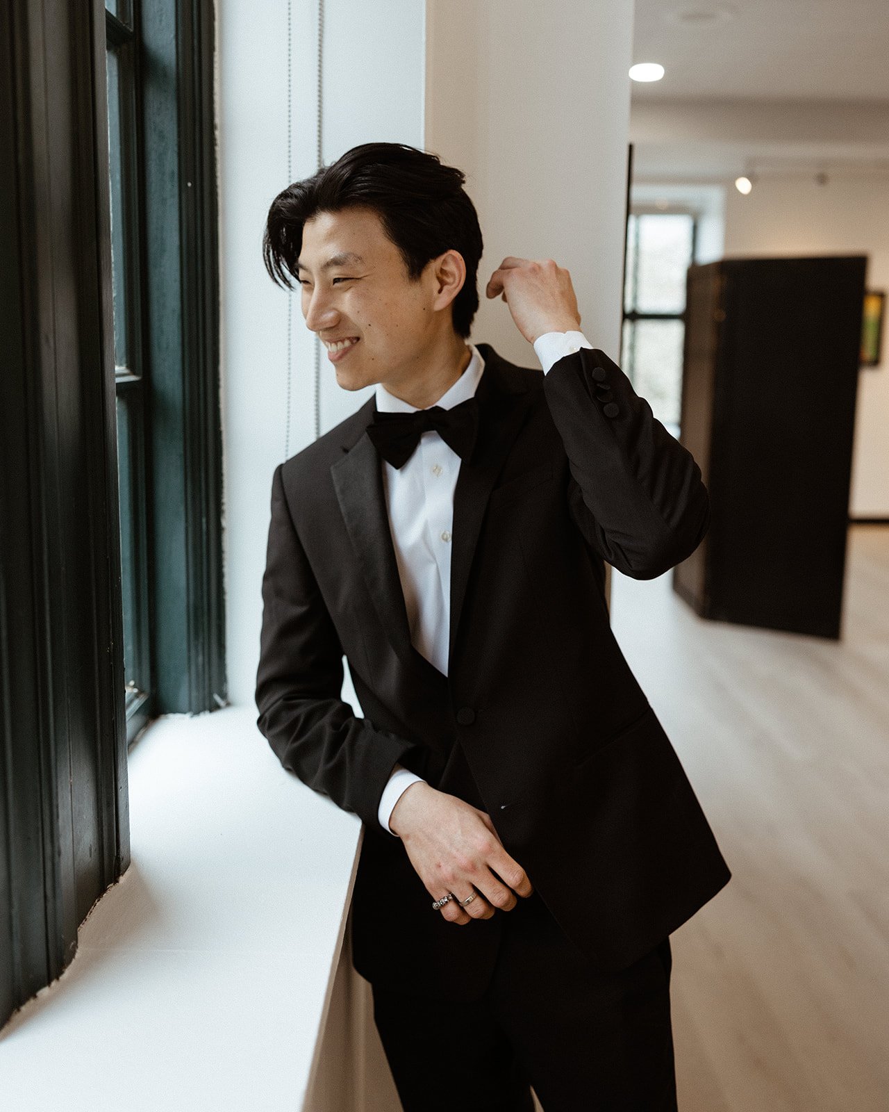 A young man dressed in a black tuxedo with a bow tie, smiling and adjusting his collar while standing near a window in a modern room.