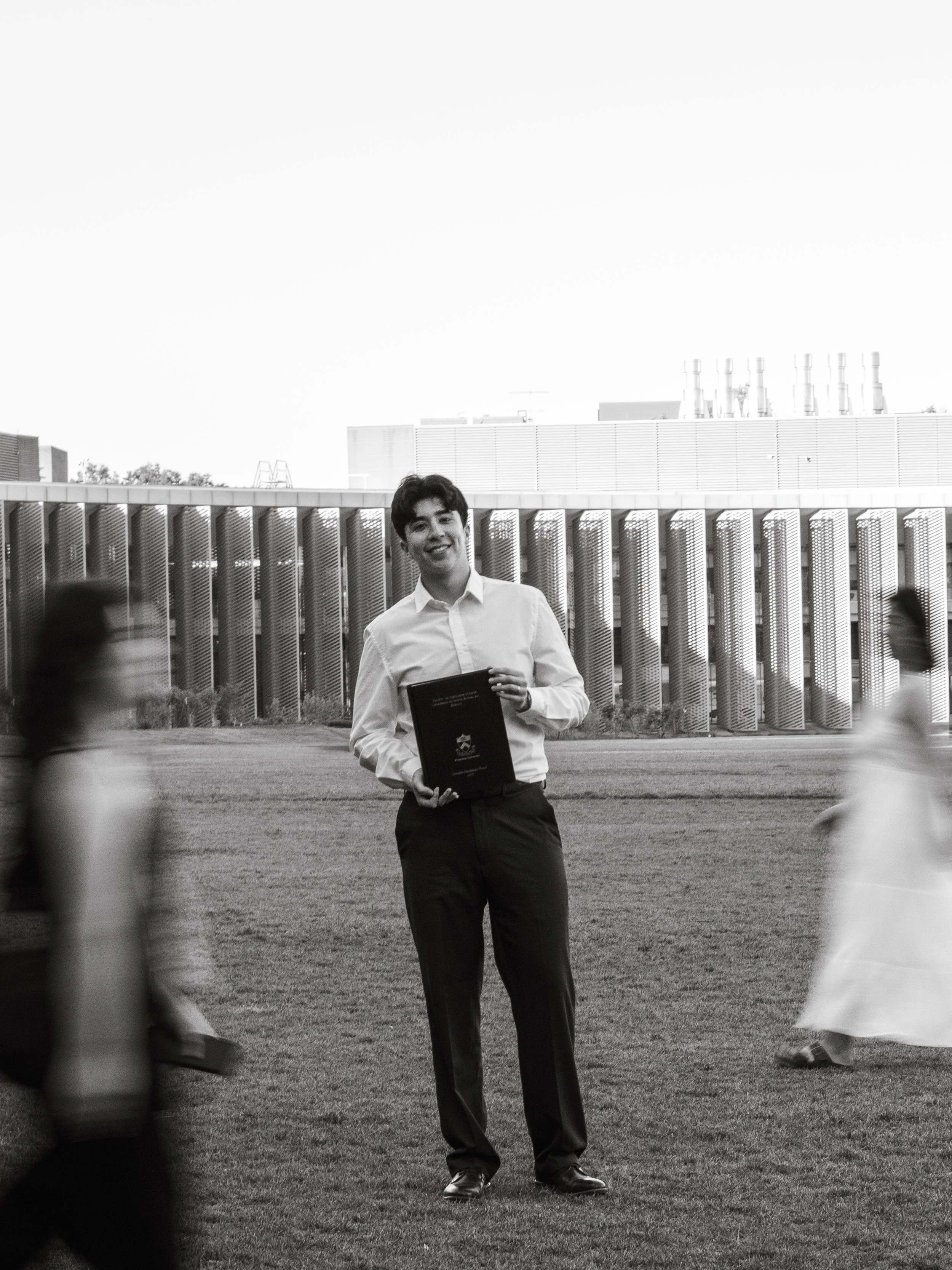 A young man in a white shirt and dark pants smiling while holding a dark folder, standing on a grassy area with a modern building with vertical metal slats behind him, and two children partially visible in the foreground.