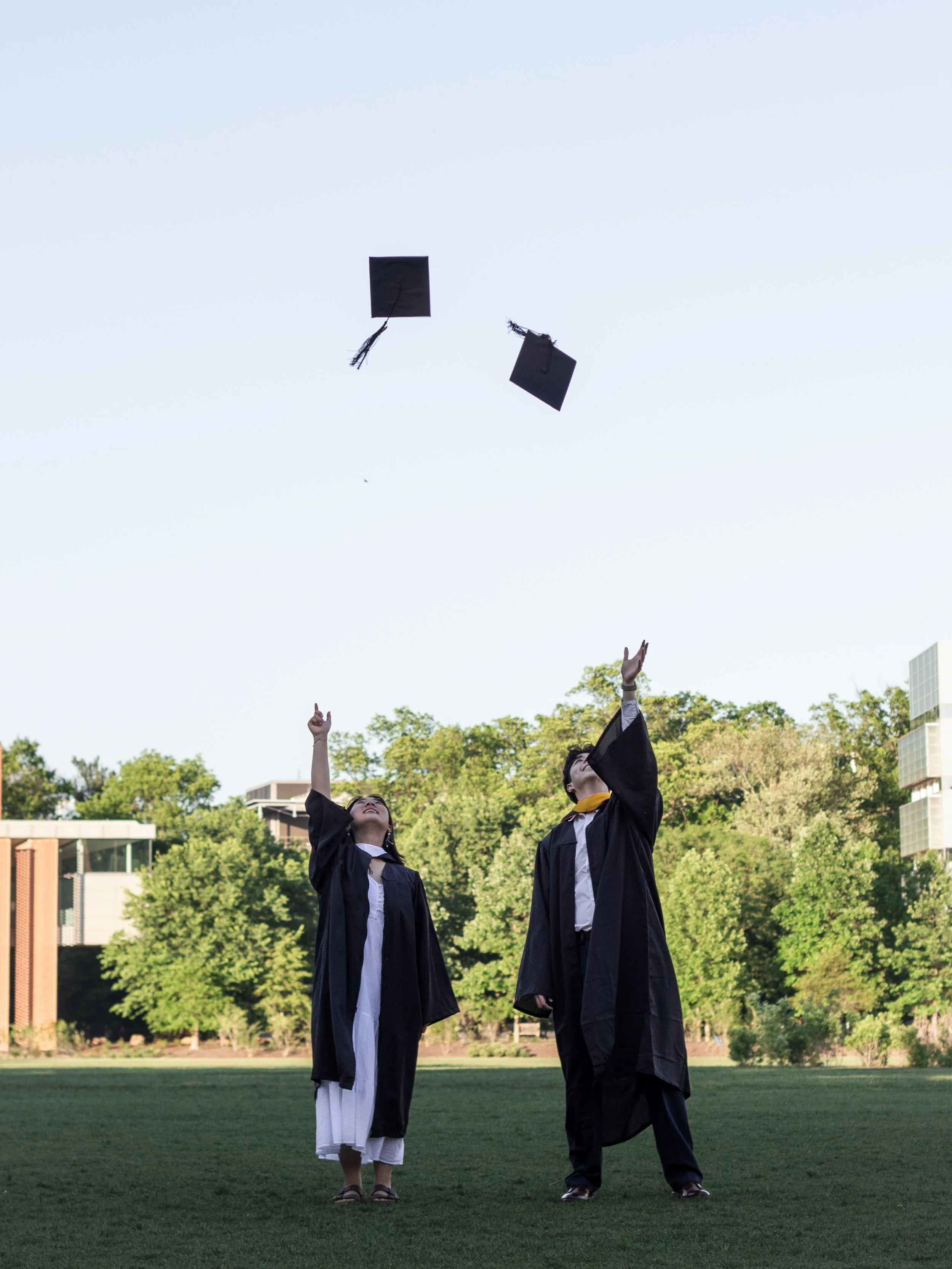 Two graduates in black gowns and caps release flying caps into the air on a grassy field with trees and buildings in the background.