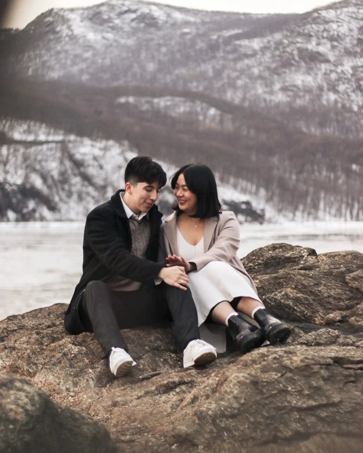 A young man and woman sitting on rocks by a body of water surrounded by mountains, sharing a tender moment and smiling at each other.