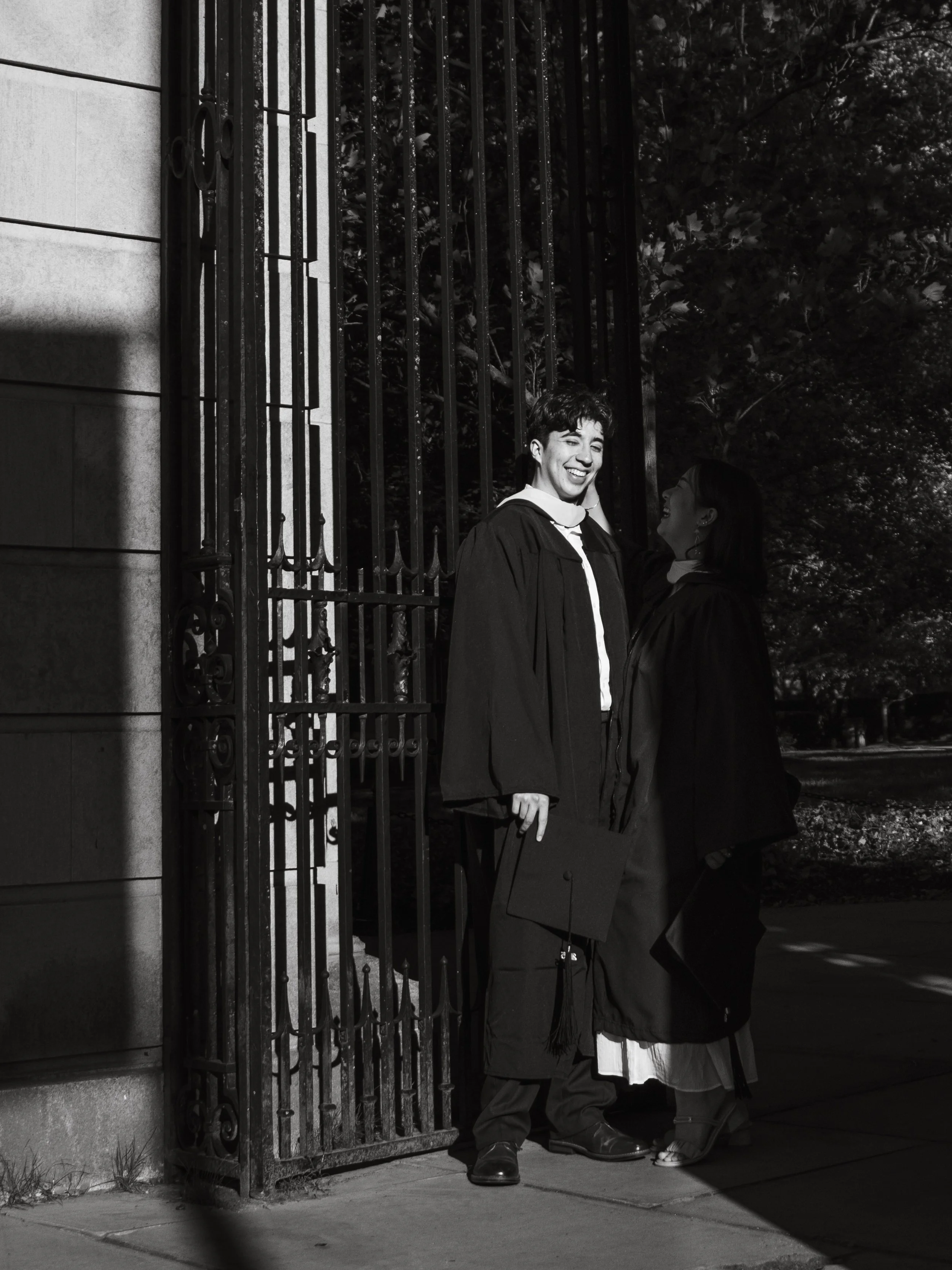 A young man and woman in graduation gowns smiling and looking at each other outside near a metal gate and trees.