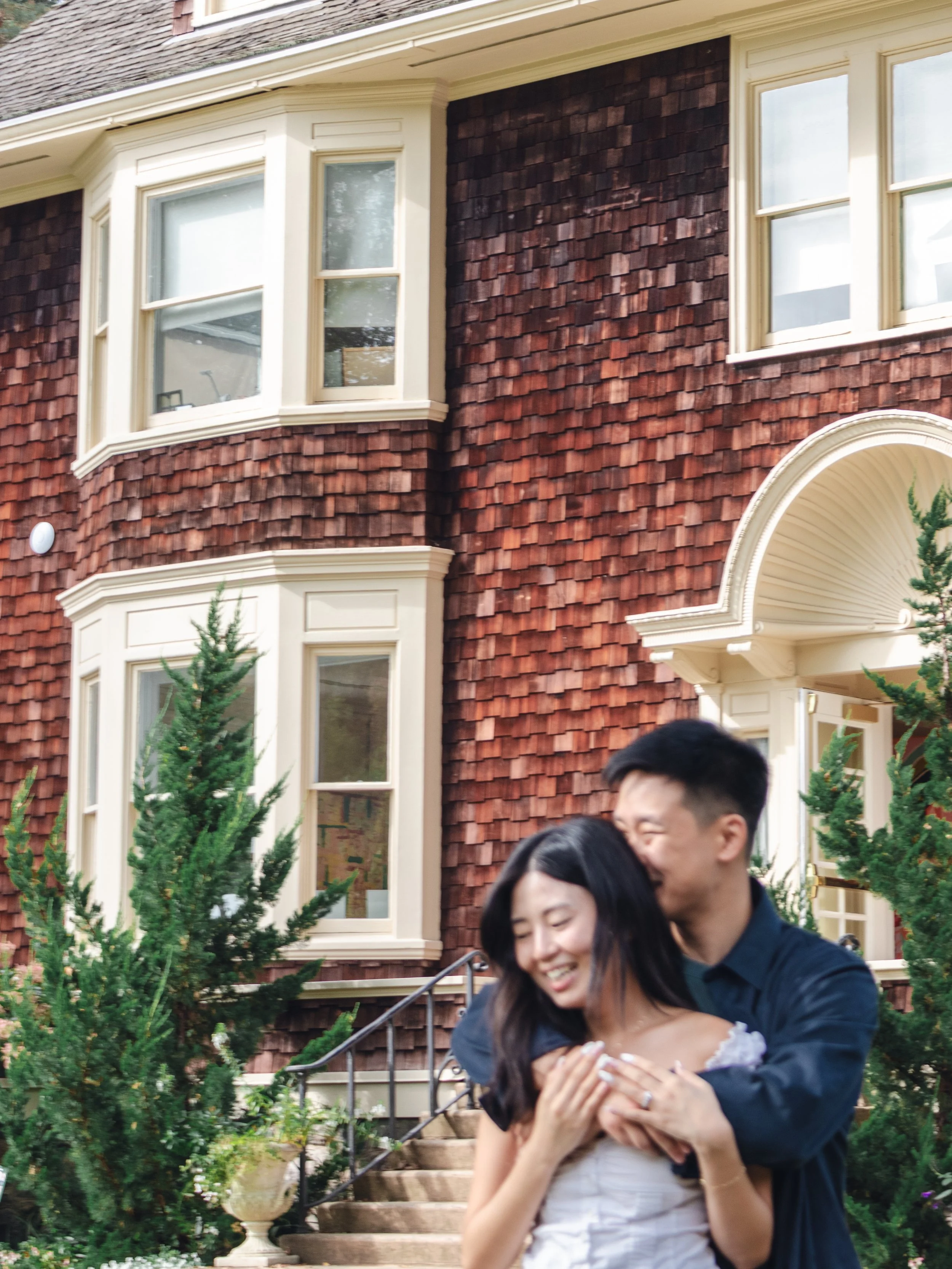 A couple is joyfully embracing outside a red-brick house with white trim and large windows, surrounded by green trees and plants.