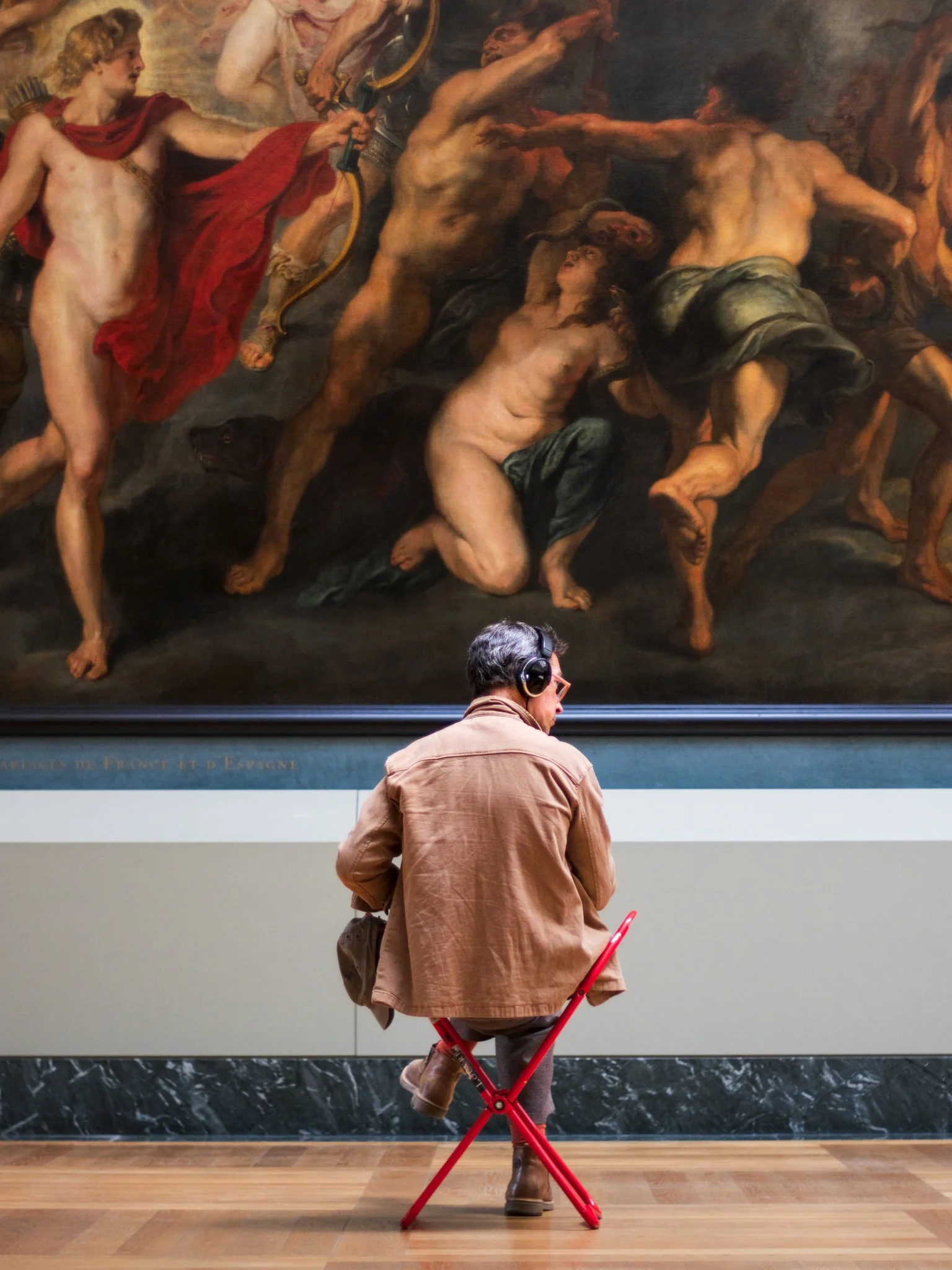 A person sitting on a red folding chair in front of a large painting in an art gallery. The person is wearing a brown jacket, jeans, brown boots, and glasses, and has headphones on. The painting in the background depicts a dramatic scene with several nude figures in dynamic poses.