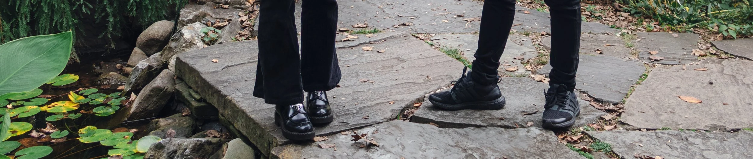 Close-up of two people's feet standing on a stone pathway next to a small pond with lily pads and large green leaves.