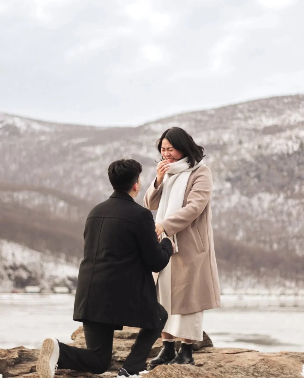 A man is kneeling and proposing to a woman by a lake with mountains in the background. The woman is smiling and holding the man's hand. Both are dressed warmly in coats, and the woman is wearing a white scarf.