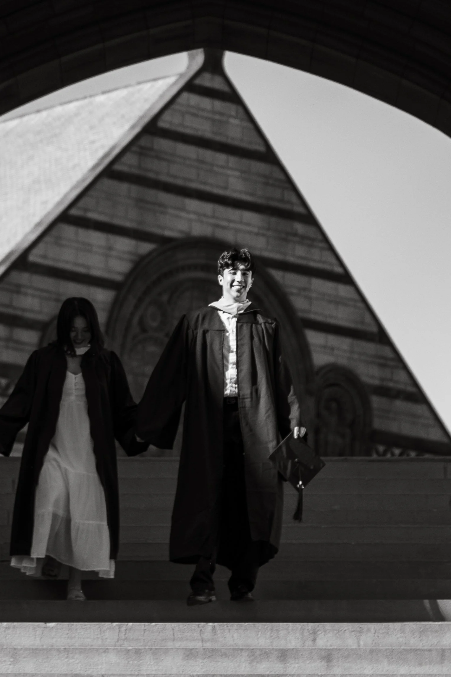Black and white photo of three students in graduation caps and gowns descending stairs outside a building with rounded architecture and a large decorative arch.