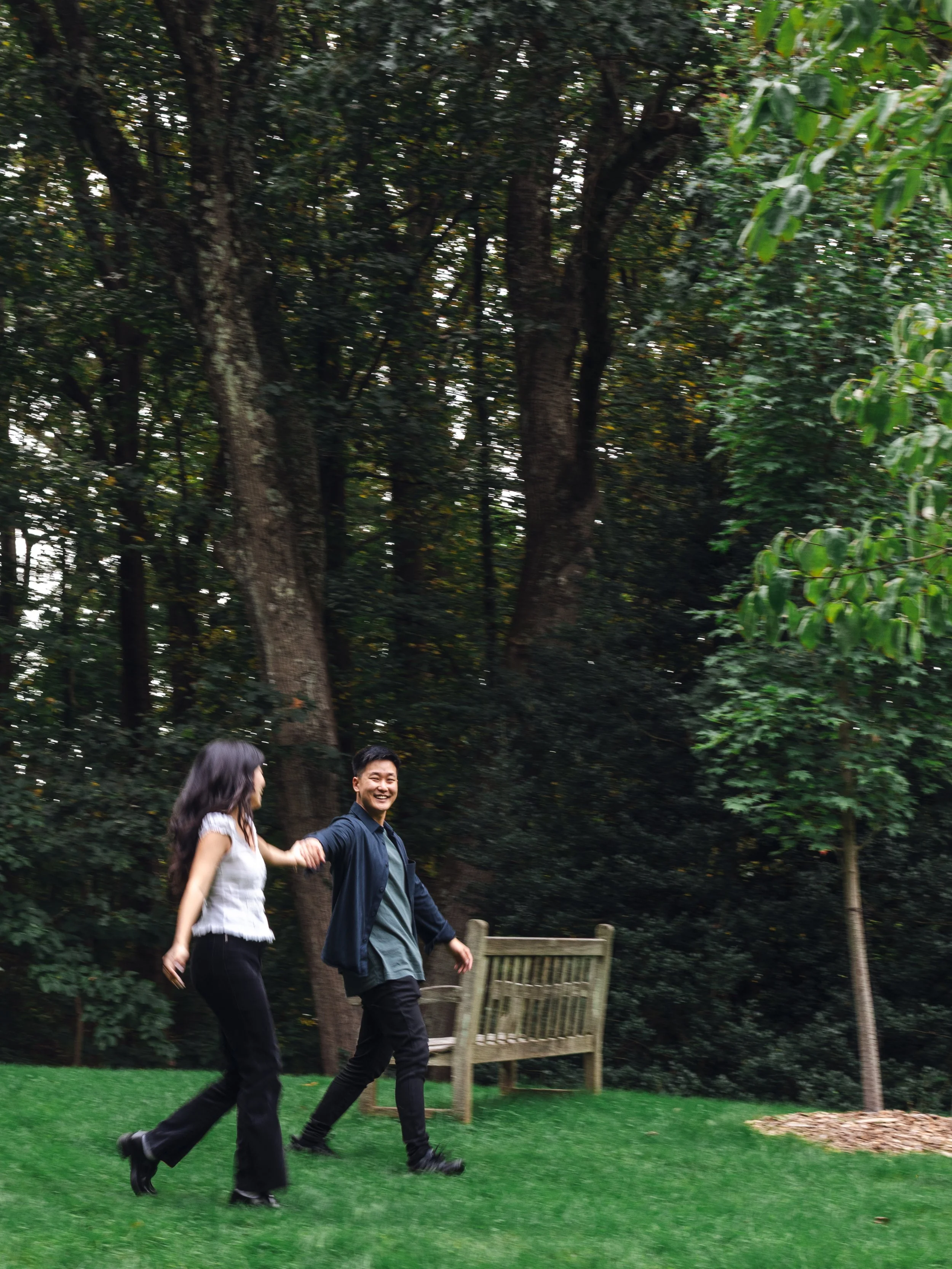 A man and a woman are walking hand in hand in a park with lush green grass and tall trees in the background. They are smiling and appear to be enjoying a walk together.
