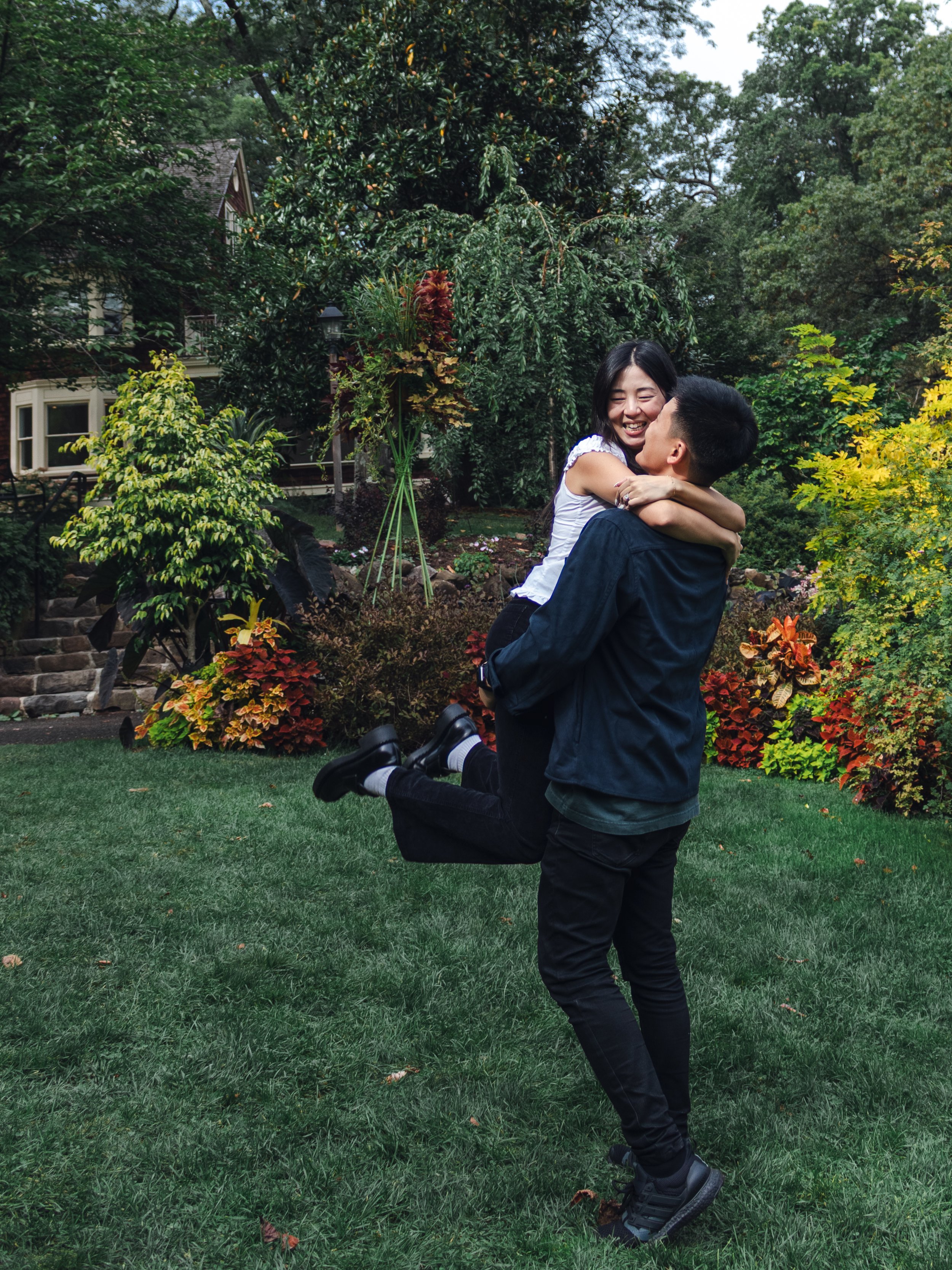 A young couple is outdoors in a garden, with the man kneeling and lifting the woman in his arms. They are smiling and appear happy, surrounded by lush green trees and colorful flowers.